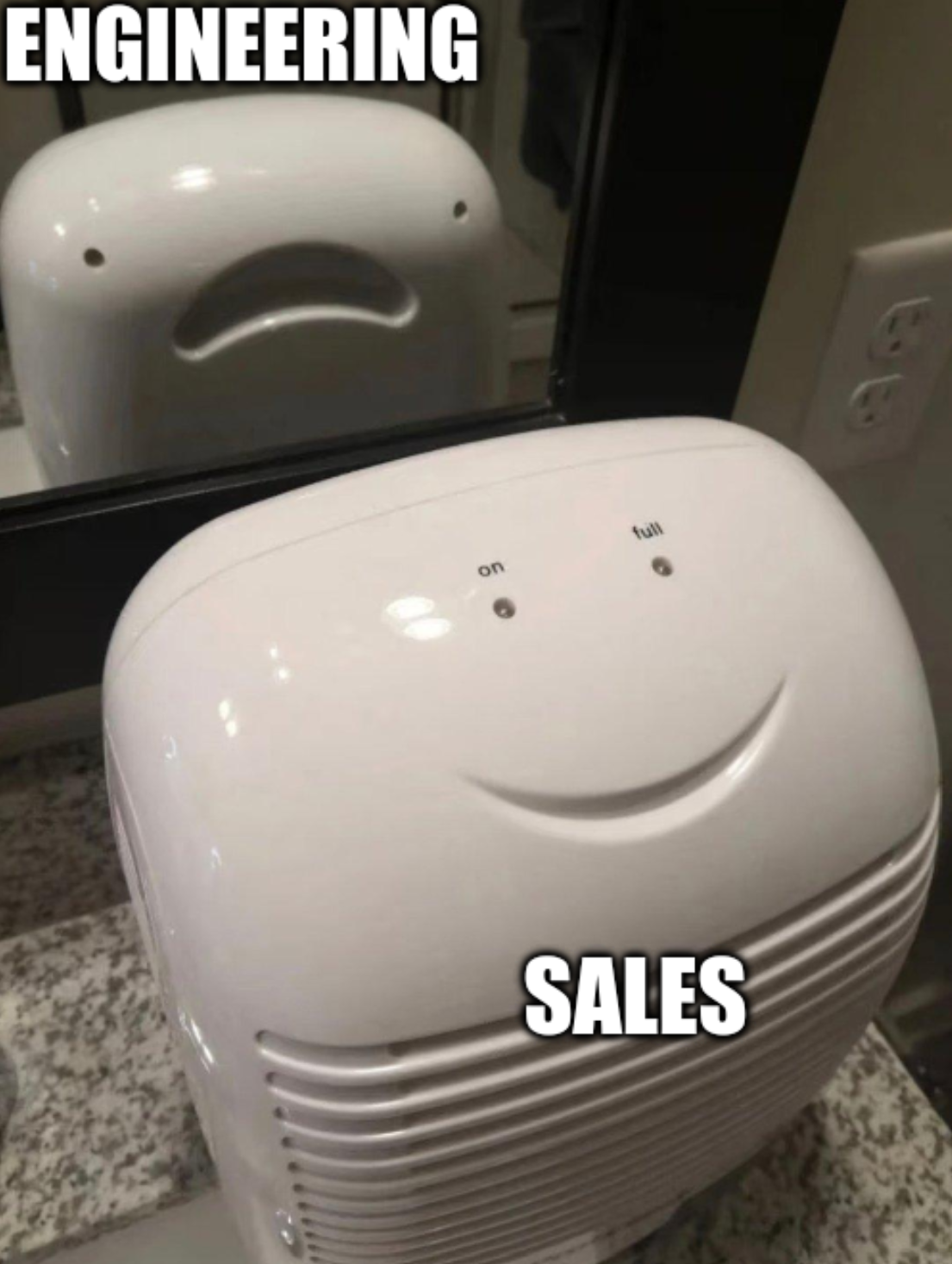 Photo of a dehumidifier in front of the mirror. The front of it is labeled "Sales" and the lights and shape make it look like a smiley face. The reflection of the back in the mirror is labeled "Engineering" and the screw holes and handle make it look like it's a terrified face.