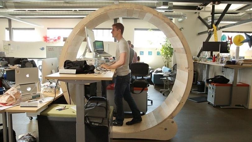 Photo of an office, presumably of a tech company. Open space, desks with laptops and screens. In the foreground a white male programmer at his desk, walking slowly inside a huge wooden hamster wheel.