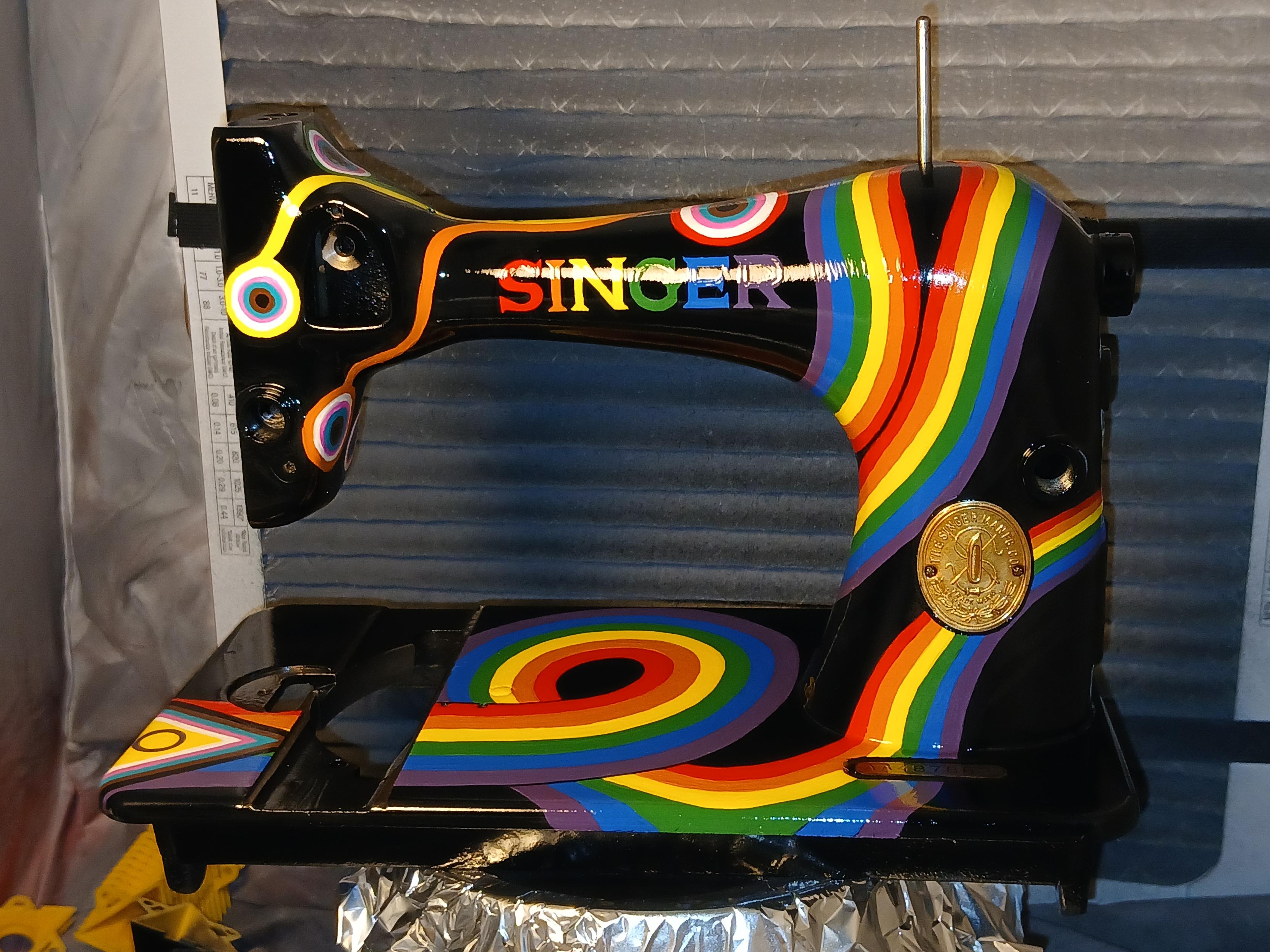 black Singer sewing machine with the progressive pride flag beginning at the lower left of the bed and wrapping around the machine terminating in the rainbow colors separated and with round baubles on the ends. white, blue, pink, brown and black form bullseyes inside. the letters of the word Singer are painted in the colors of the rainbow.