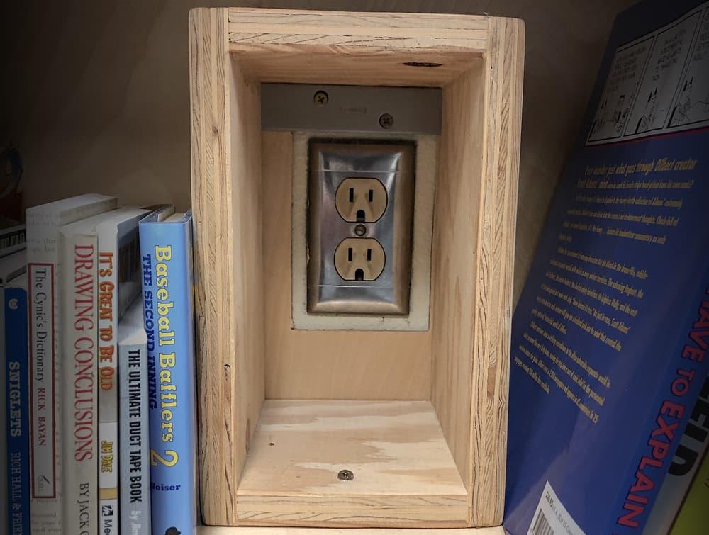 A wall outlet peeking through a makeshift wooden box that has been screwed into place on a bookshelf. Books line the shelf on each side of it.