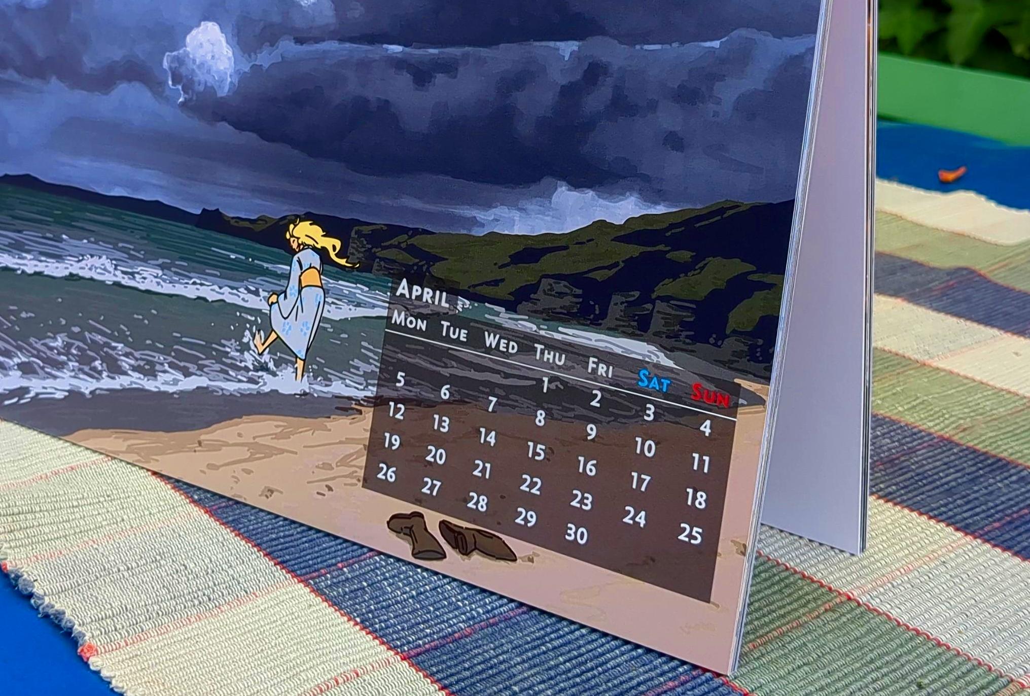 A close-up photo of an A4 calendar resting on a table. The month of April is visible, with a little day calendar showing the day of week and each day of the month of April 2027. The calendar page features a painting of a beach, with heavy clouds in the distance. A woman is playing in the shallow water.