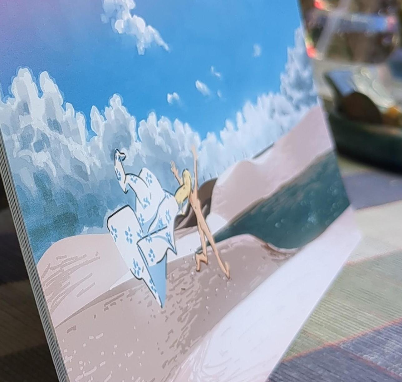 A close-up photo of an A4 calendar resting on a table. The calendar page features a painting of a sandy desert, with light clouds at the horizon. A woman is shown having thrown off her kimono and running towards a lake.