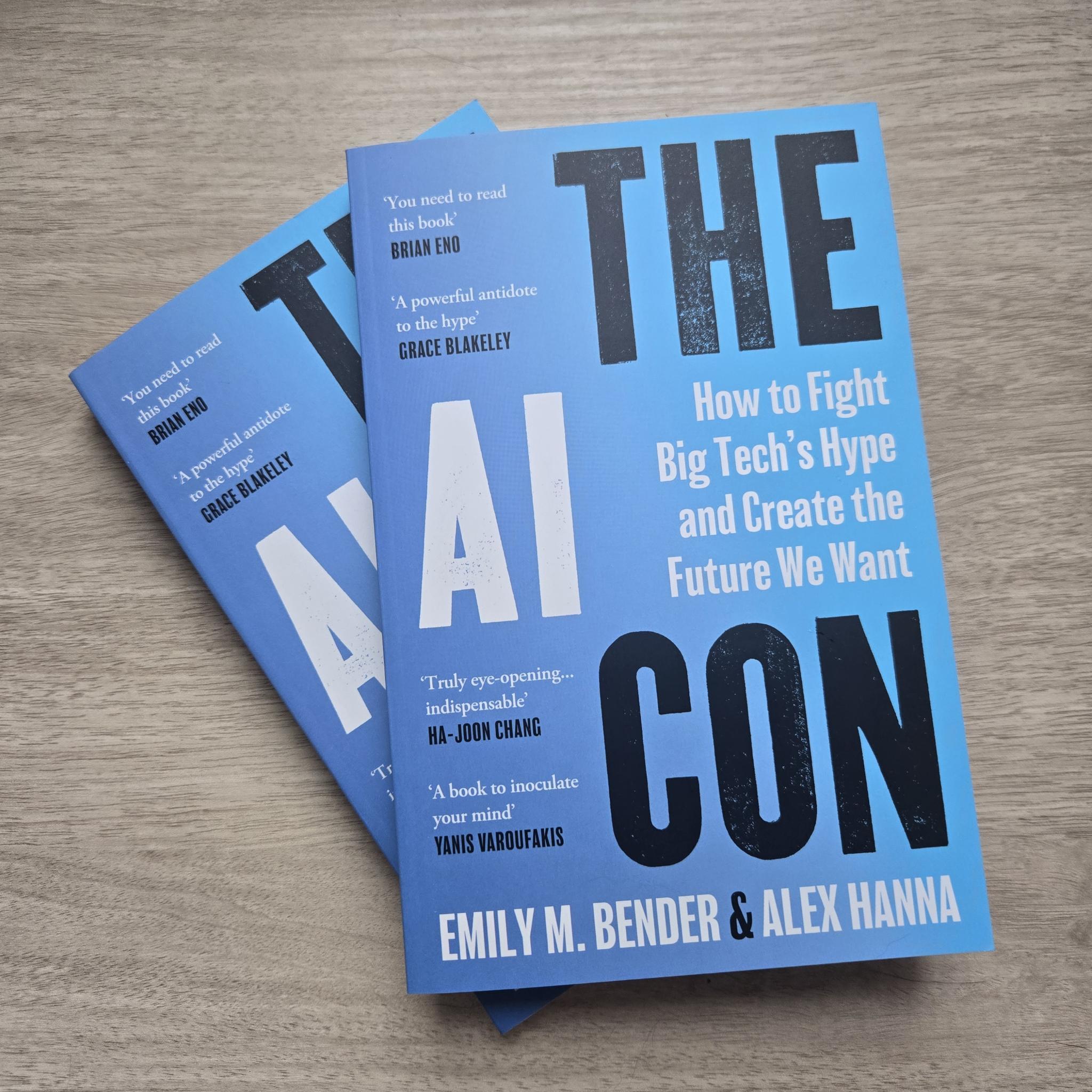 Two paperback copies of The AI Con stacked at an angle, against a light wood desk. The title is rendered in large letter-press font, surround the subtitle in smaller letters, "How to Fight Big Tech's Hype and Create the Future We Want". Author names in all caps at the bottom: "Emily M. Bender & Alex Hanna". In the upper left are two blurbs: "You need to read this book" (Brian Eno) and "A powerful antidote to the hype" (Grace Blakeley). In the lower left are two more: "Truly eye-opening ... indispensable" (Ha-Joon Chang) and "A book to inoculate your mind" (Yanis Varoufakis). The background of the cover is a blue gradient, going from almost periwinkle on the left to a bright azure on the right. The lettering is all either black or white.
