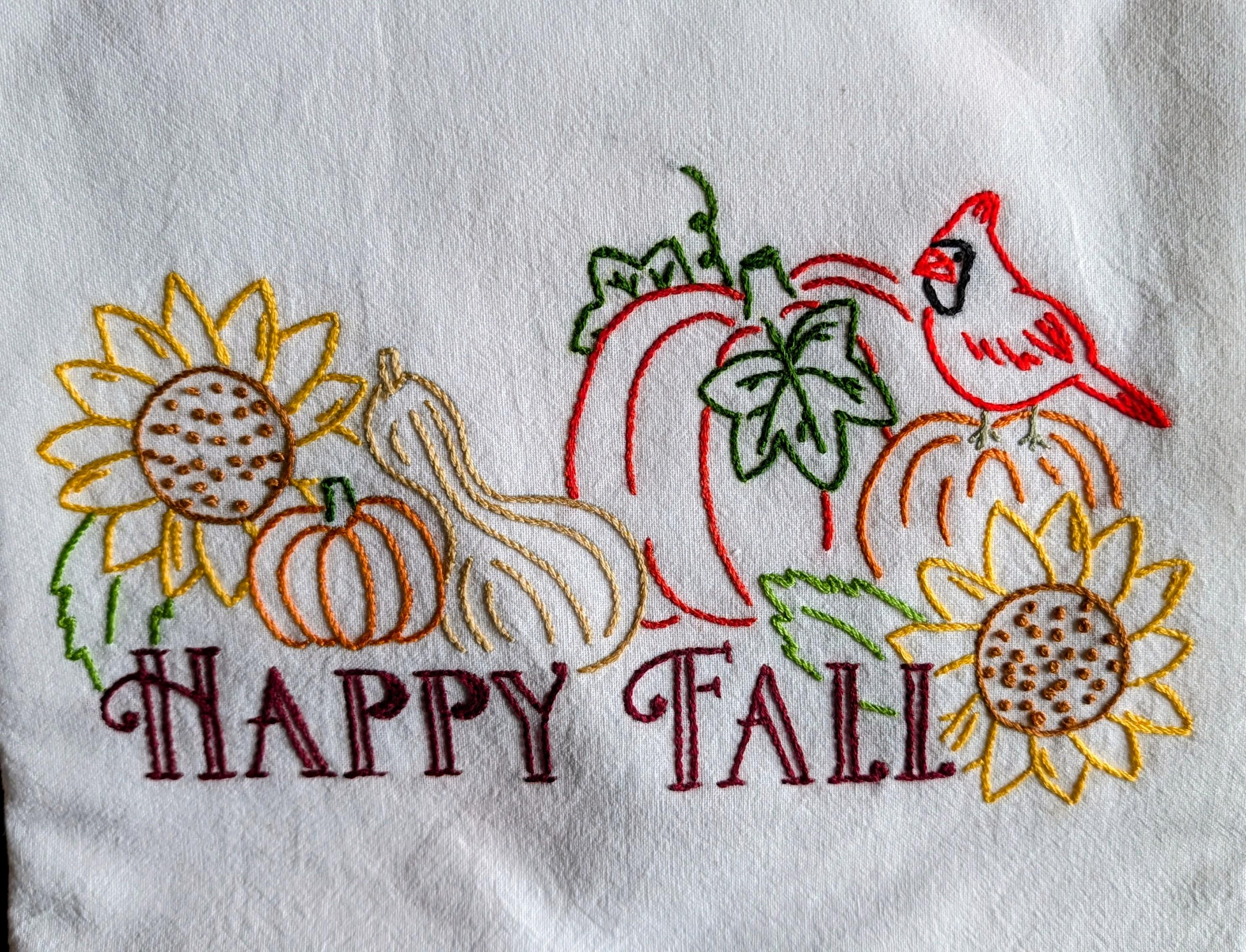 A white flour sack dishtowel embroidered with a Happy Fall design. There are sunflowers, pumpkins, a gourd and a red cardinal in the design.