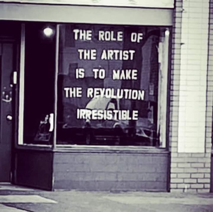 A black-and-white photograph of a storefront window featuring a large poster with bold text: “The role of the artist is to make the revolution irresistible".