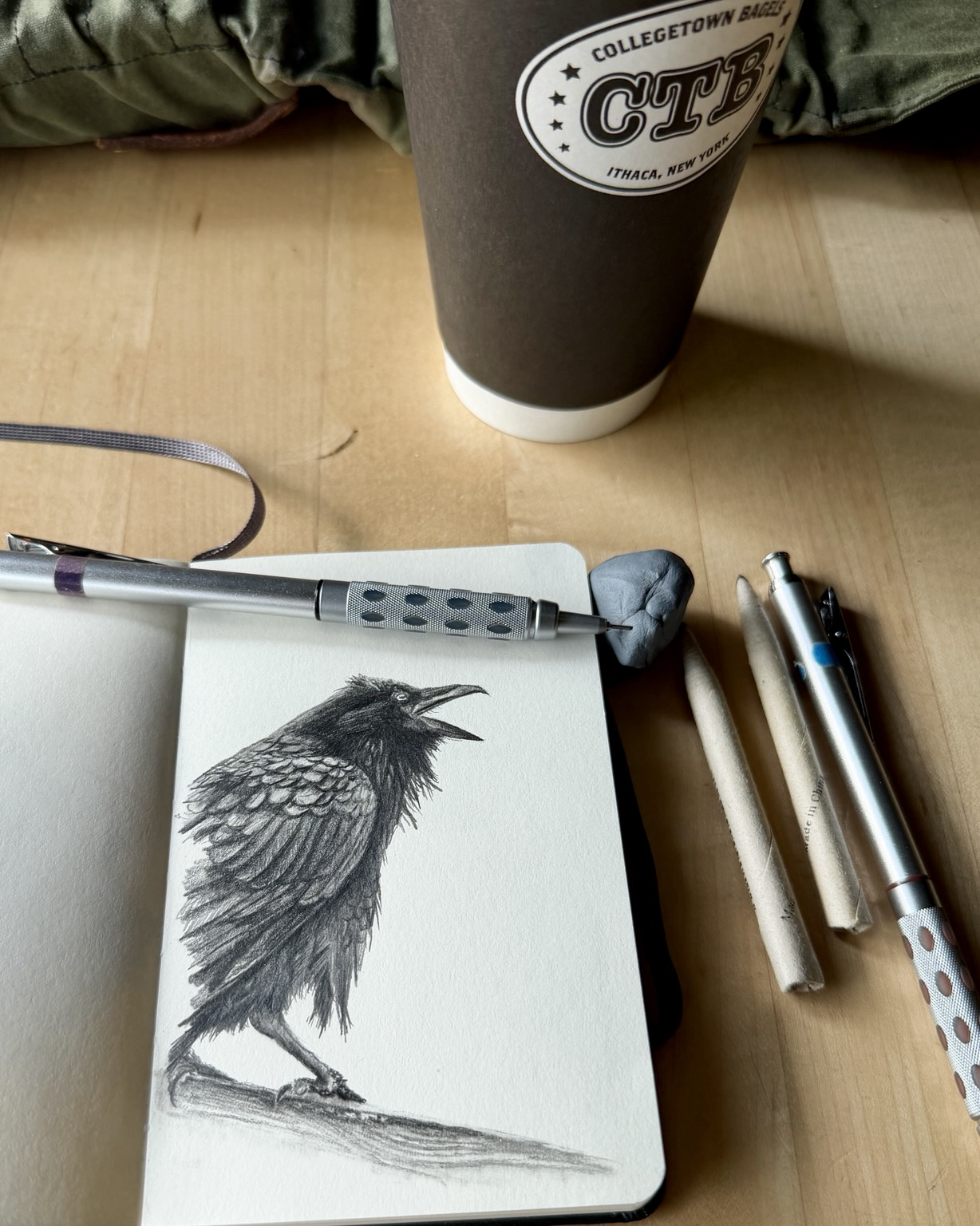 Graphite sketch of a raven mid call, beak open, feathers rendered in fine detailed strokes with dramatic tonal contrast. The bird stands on a branch, one leg lifted, set against a mostly blank cream page. A mechanical pencil, kneaded eraser, and coffee cup rest beside the sketchbook on a wooden table.