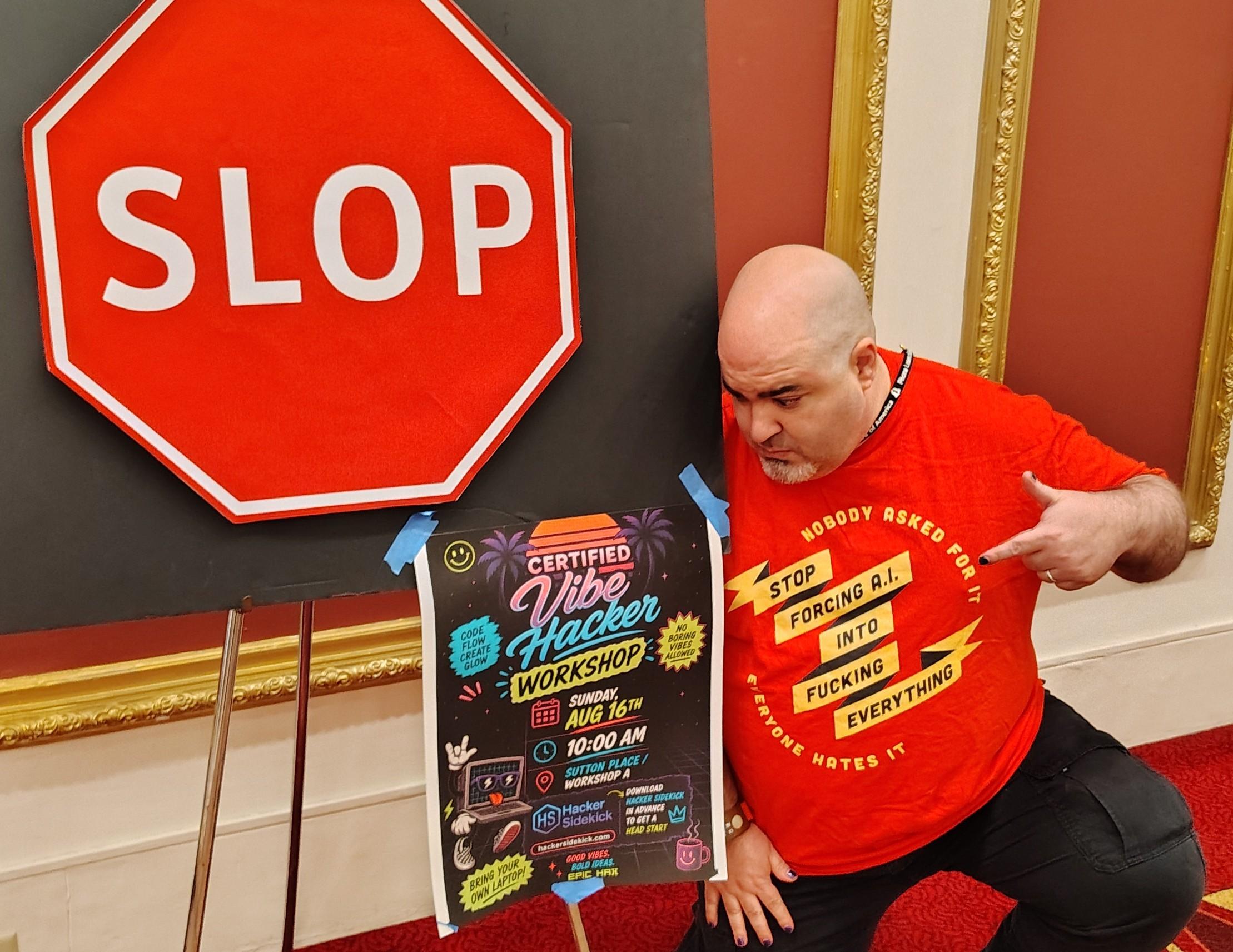 Photo of the Undersigned crouching near a poster for a "vibe hacking" workshop which has been enragingly posted right by an anti-A.I. "Slop" sign (a hexagonal stop sign with one letter changed.) The Undersigned is flashing at the poster and pointing at the T-shirt he is wearing, which reads "Stop forcing A.I. into fucking everything. Nobody asked for it. Everyone hates it."