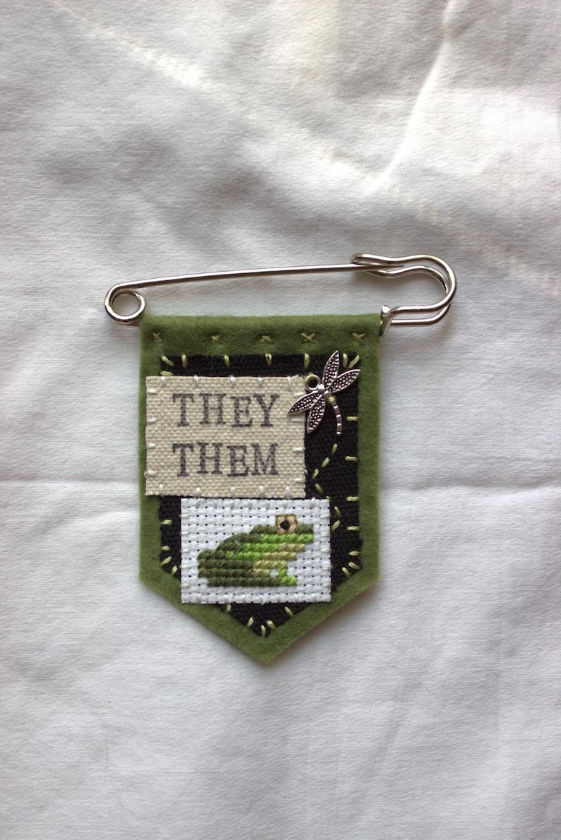 A green felt pronoun pin saying THEY THEM. it also has a cross stitch frog and a little dragon charm