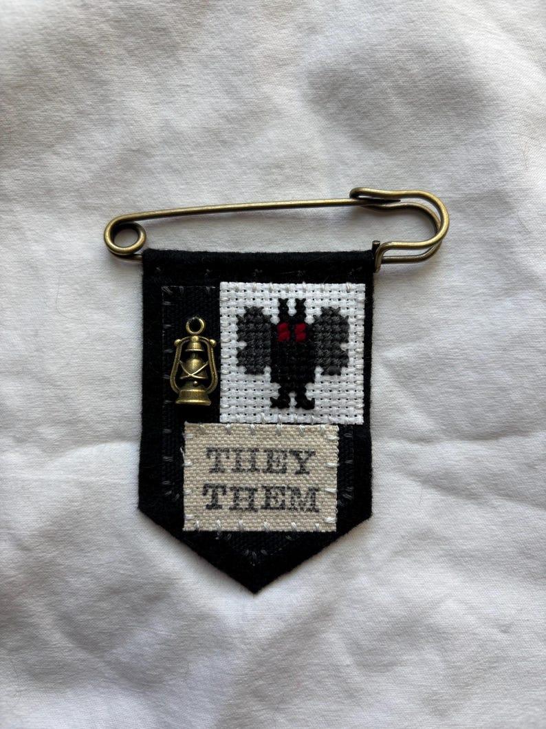 A pronoun pin with Mothman cross stitched on it and a small lantern charm. It also says THEY THEM