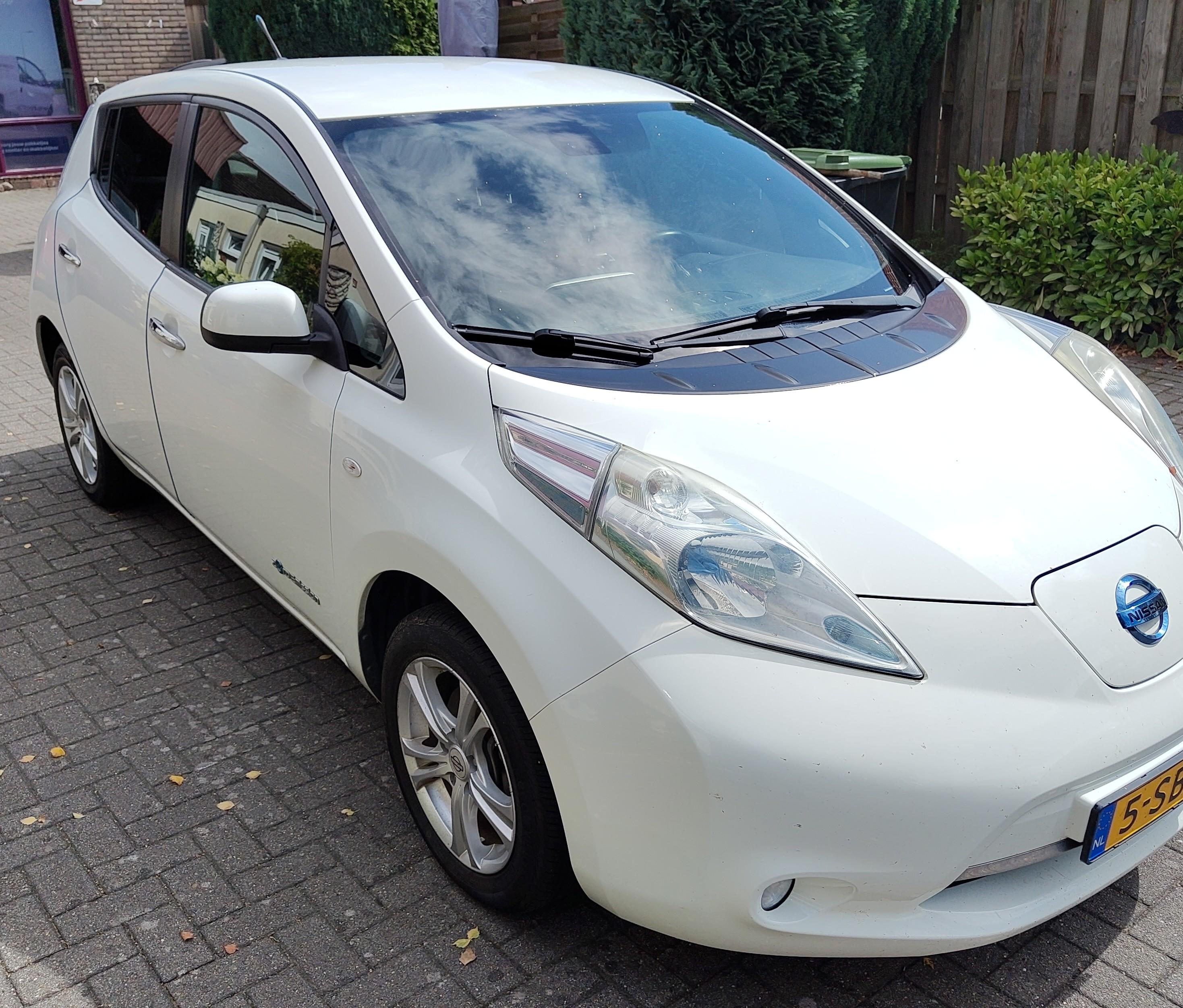 A white Nissan Leaf gen 1 sitting on a brick patio with some bushes and garden fence. It has blue door protectors giving them a deep blue edge, and a yellow dutch license plate