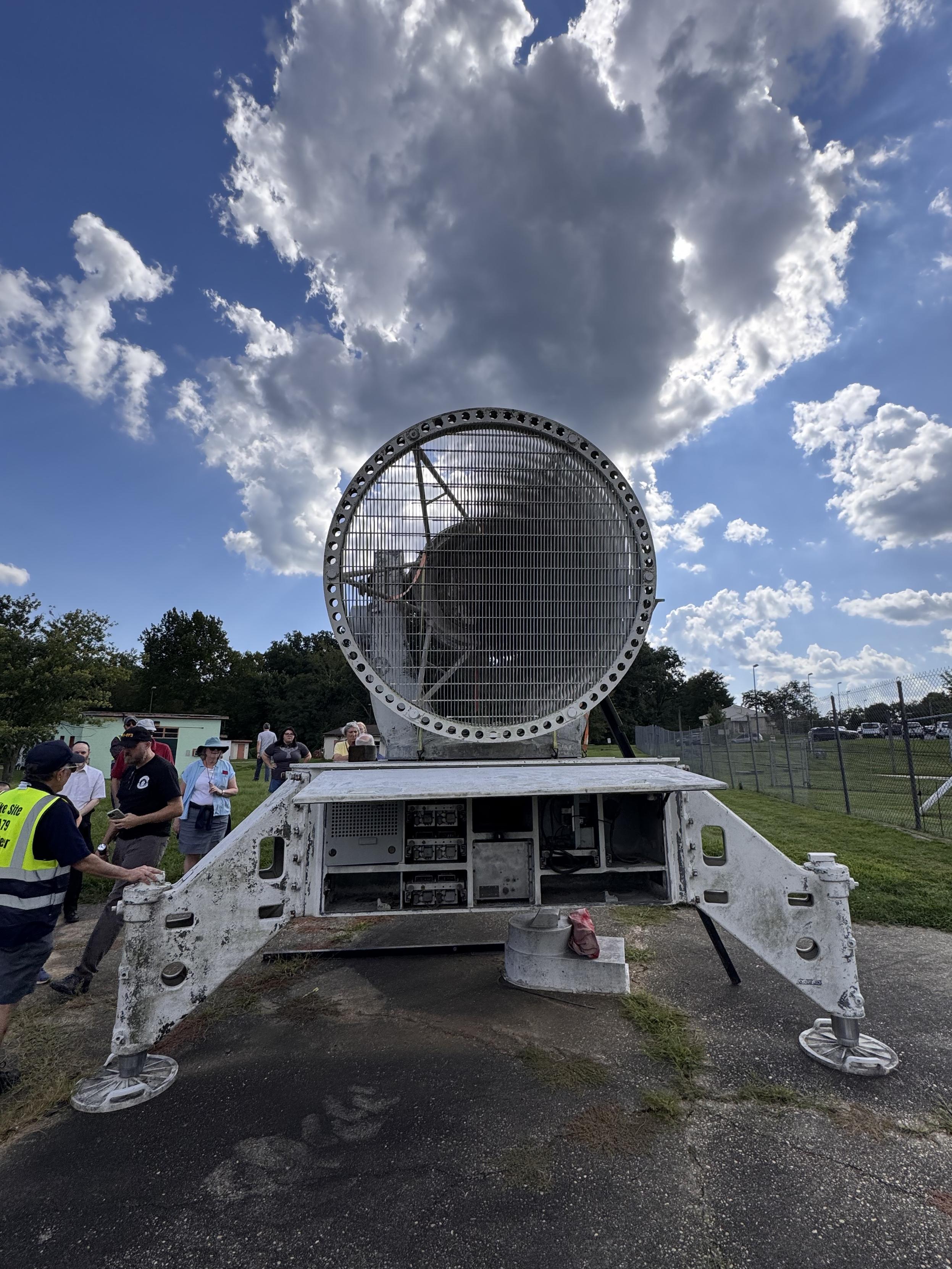 A head-on view of a decommissioned directional radar device. It stands on metal legs on an asphalt pad, viewed against a cloudy blue sky. The metal grating of the directional “dish” is cut with a Fresnel pattern, providing the directionality.