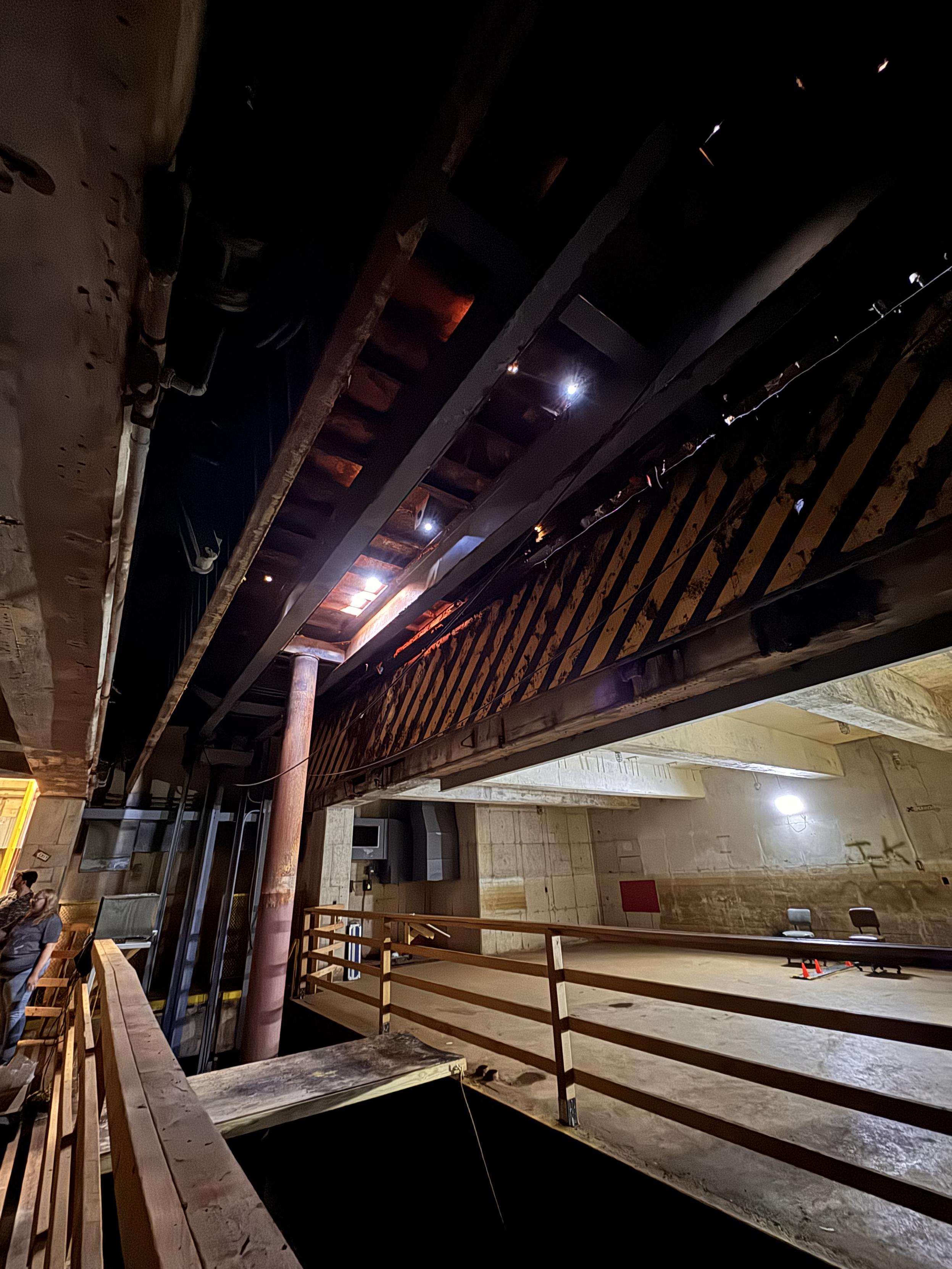 A view from below the raised elevator of an underground Nike missile magazine, showing the metal piston holding it up and the water-filled pit below. Light peeks through several rusted holes in the metal. Black and yellow stripes adorn the doors that would fold up when the elevator was lowered.