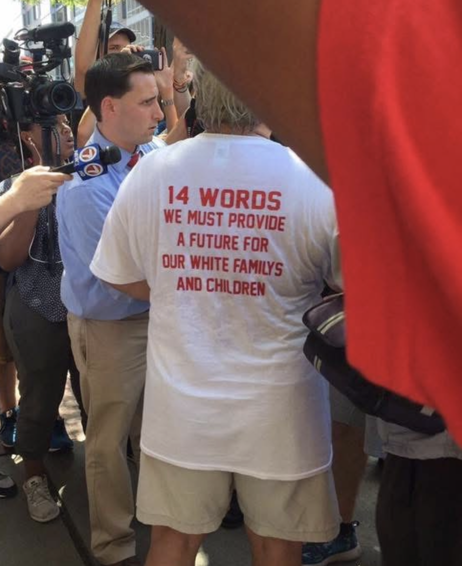 A photograph captures a group of people gathered closely together. In the foreground on the right, a large, blurry red shape partially obscures the view. Centered in the frame, a person seen from behind wears a white t-shir t-shirt with red text that reads, "14 WORDS WE MUST PROVIDE A FUTURE FOR OUR WHITE FAMILYS AND CHILDREN". To the left, a person in a light blue shirt and khaki pants is positioned near a microphone with a "FOX 5" logo.