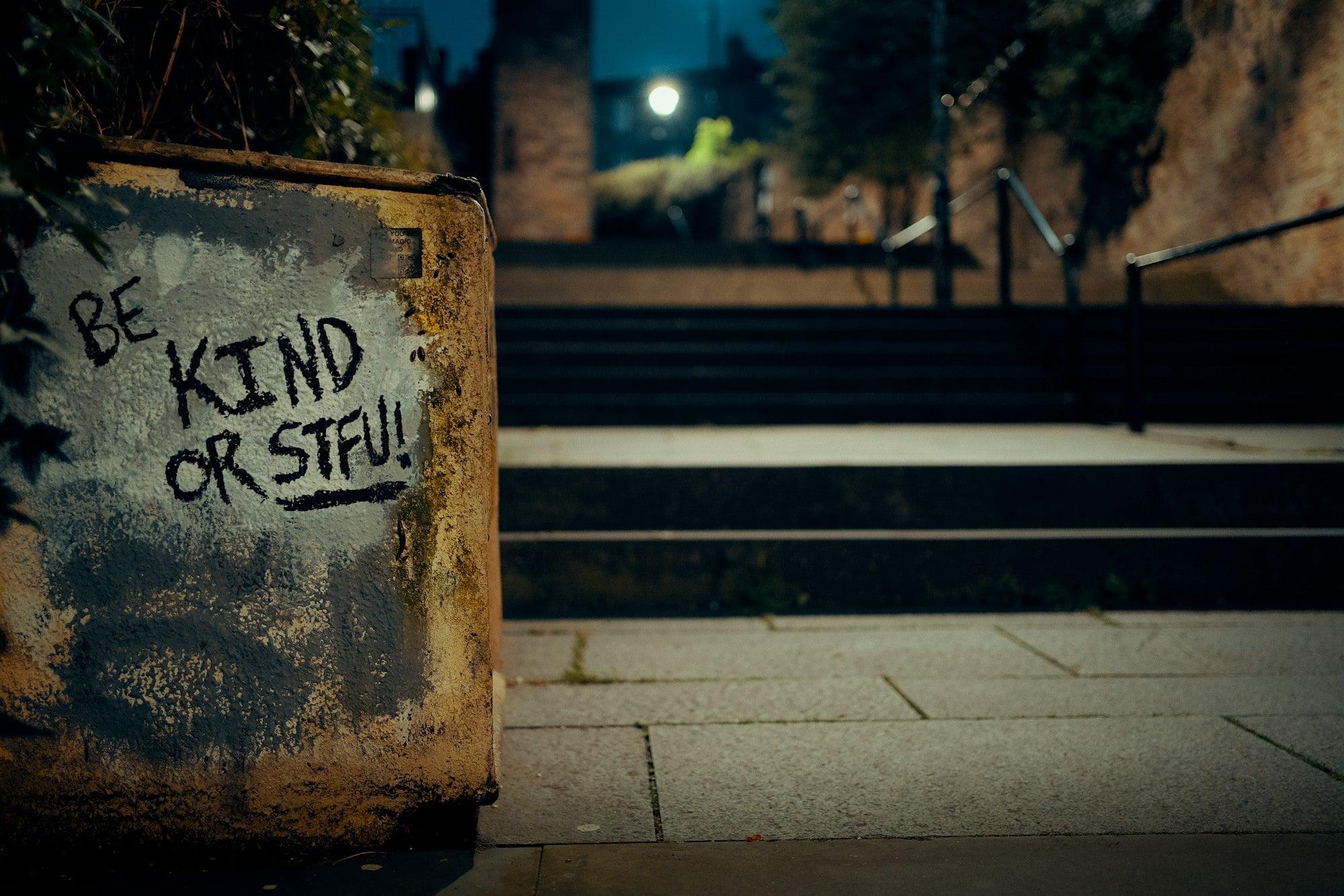 A weathered concrete block in the foreground displays graffiti reading "BE KIND OR STFU!" in black marker. In the background, a set of stairs leads upward under a dimly lit night sky.