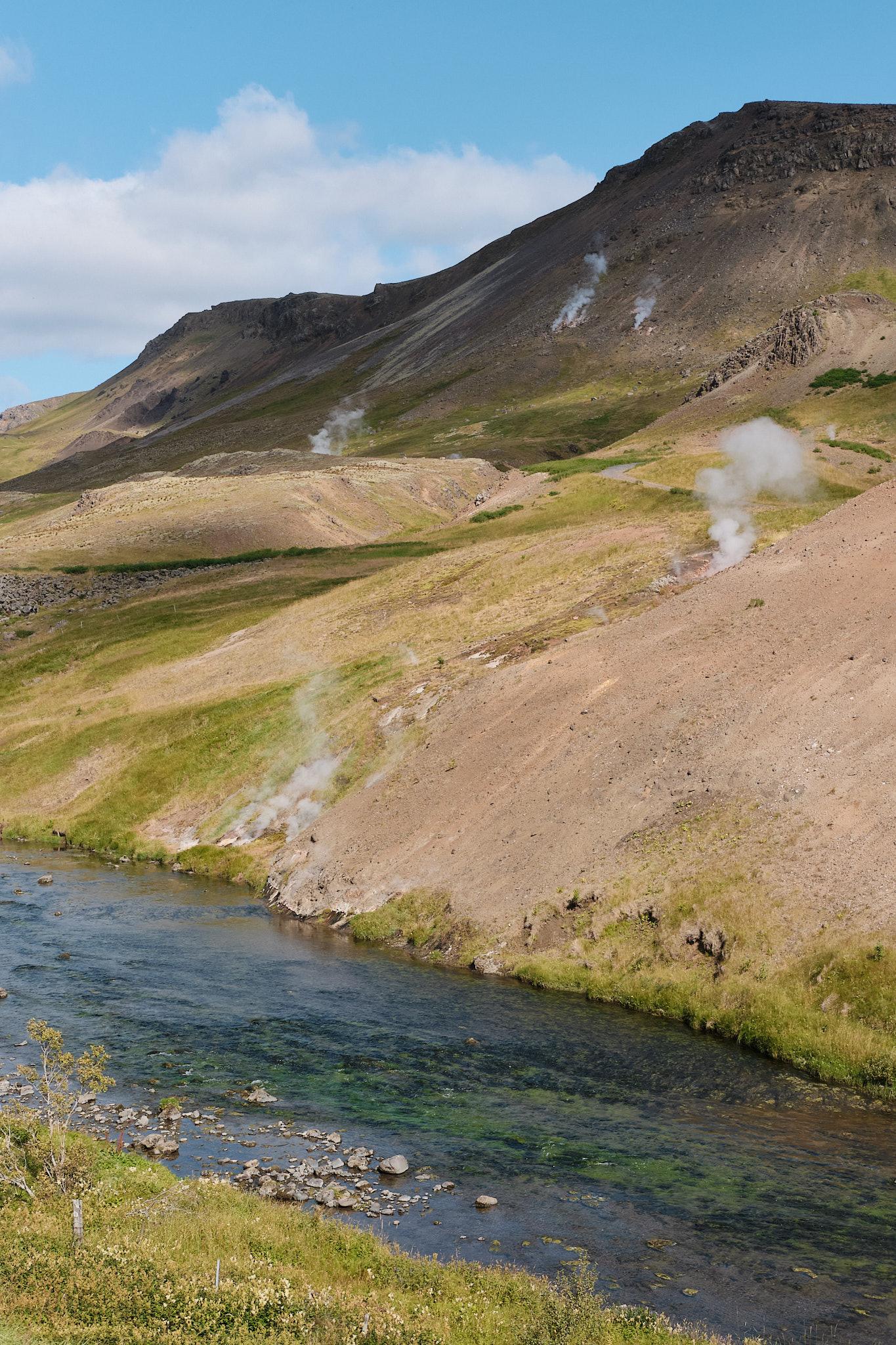 A river runs past a mountainside. Steam rises here and there throughout the landscape.
