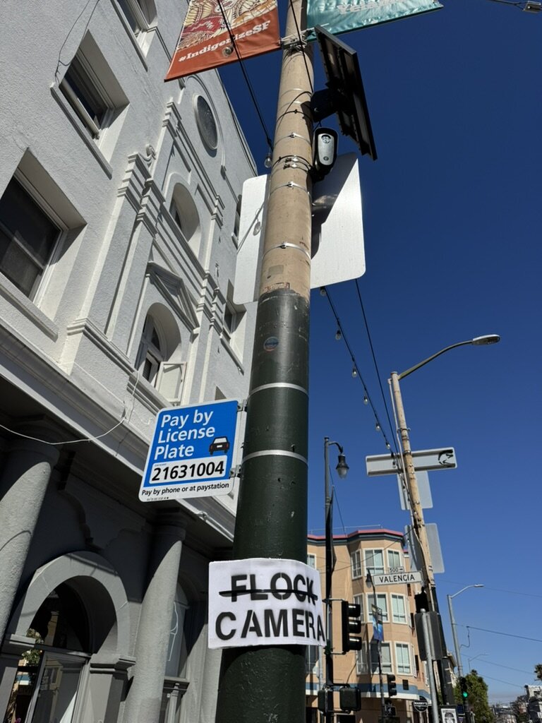 A lightpost on street corner. Someone has posted a cardboard sign around the base that says "Flock Camera". At the top of the lightpost there is a solar panel and Flock camera. The sign is crudely made using a stencil.