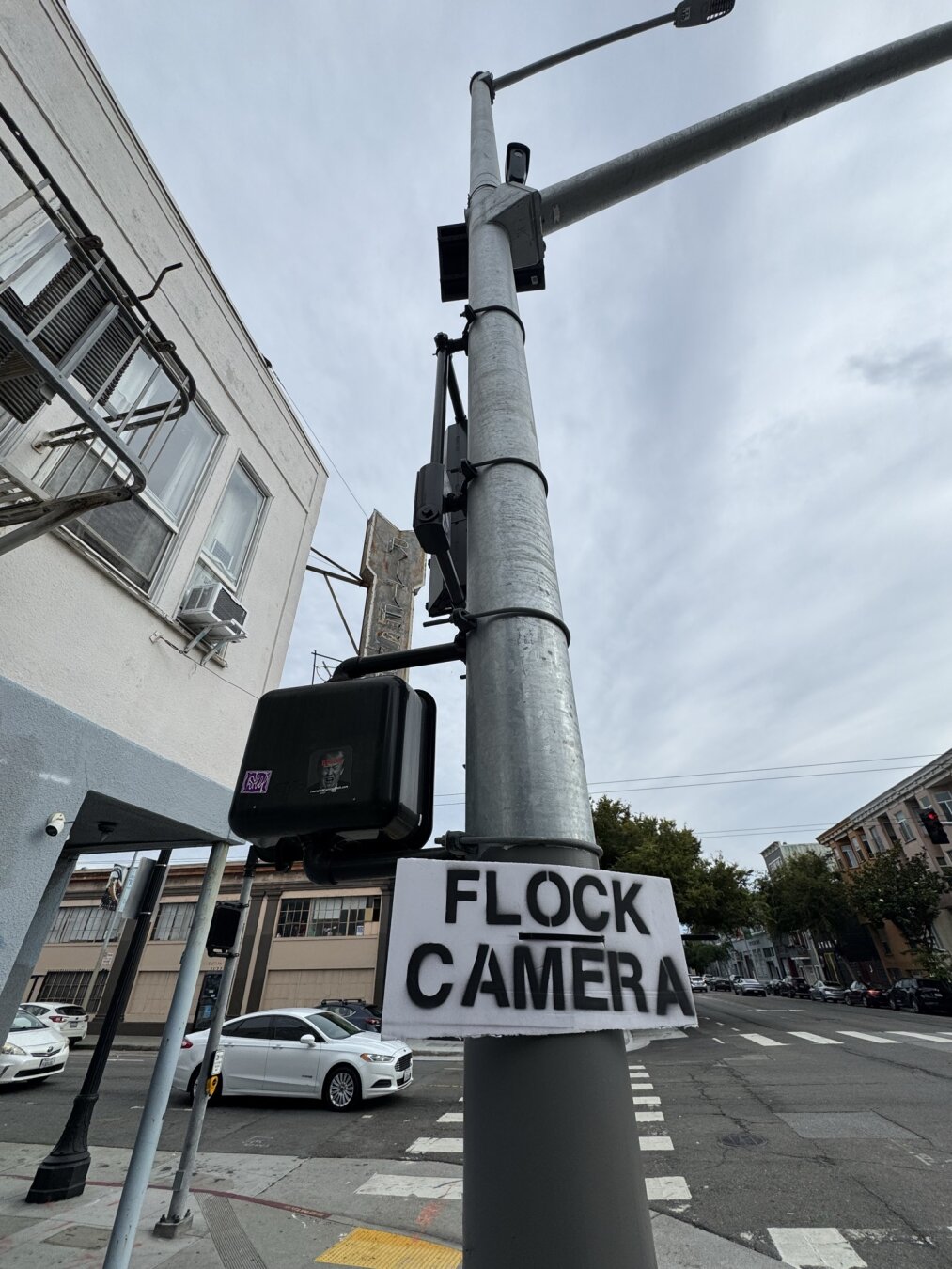 A lightpost on street corner. Someone has posted a cardboard sign around the base that says "Flock Camera". At the top of the lightpost there is a solar panel and Flock camera. The sign is crudely made using a stencil. This is similar to the other photo, but a different street corner.