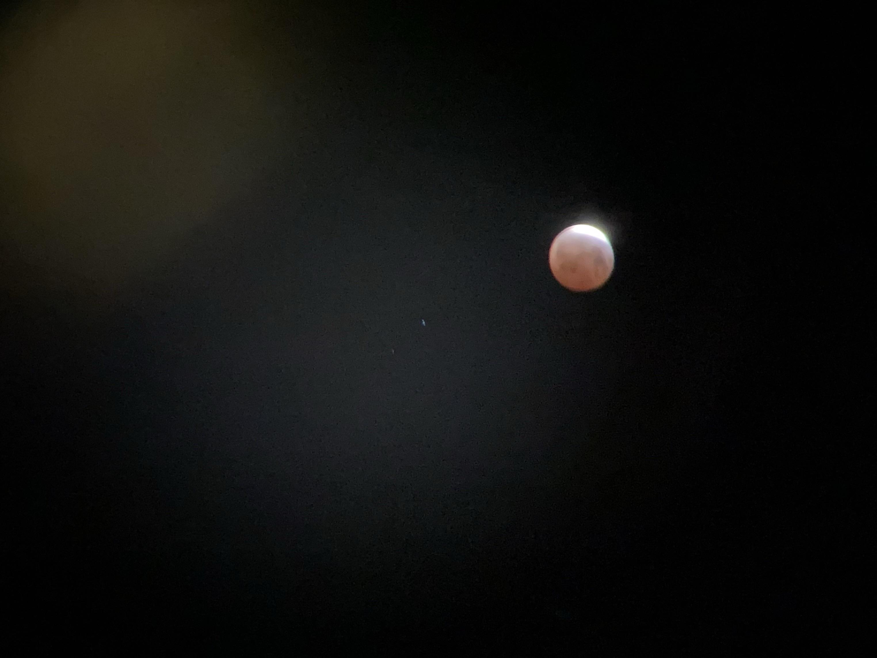 The moon during an almost total eclipse. The sky is black. The moon has a sliver of white at the top right, and is otherwise red with the earth's shadow. There's a star or planet close to the moon, and a couple of light artifacts from the camera lens and/or binoculars, making a light orange blur at the top left and a bluish blur just below it to the right.