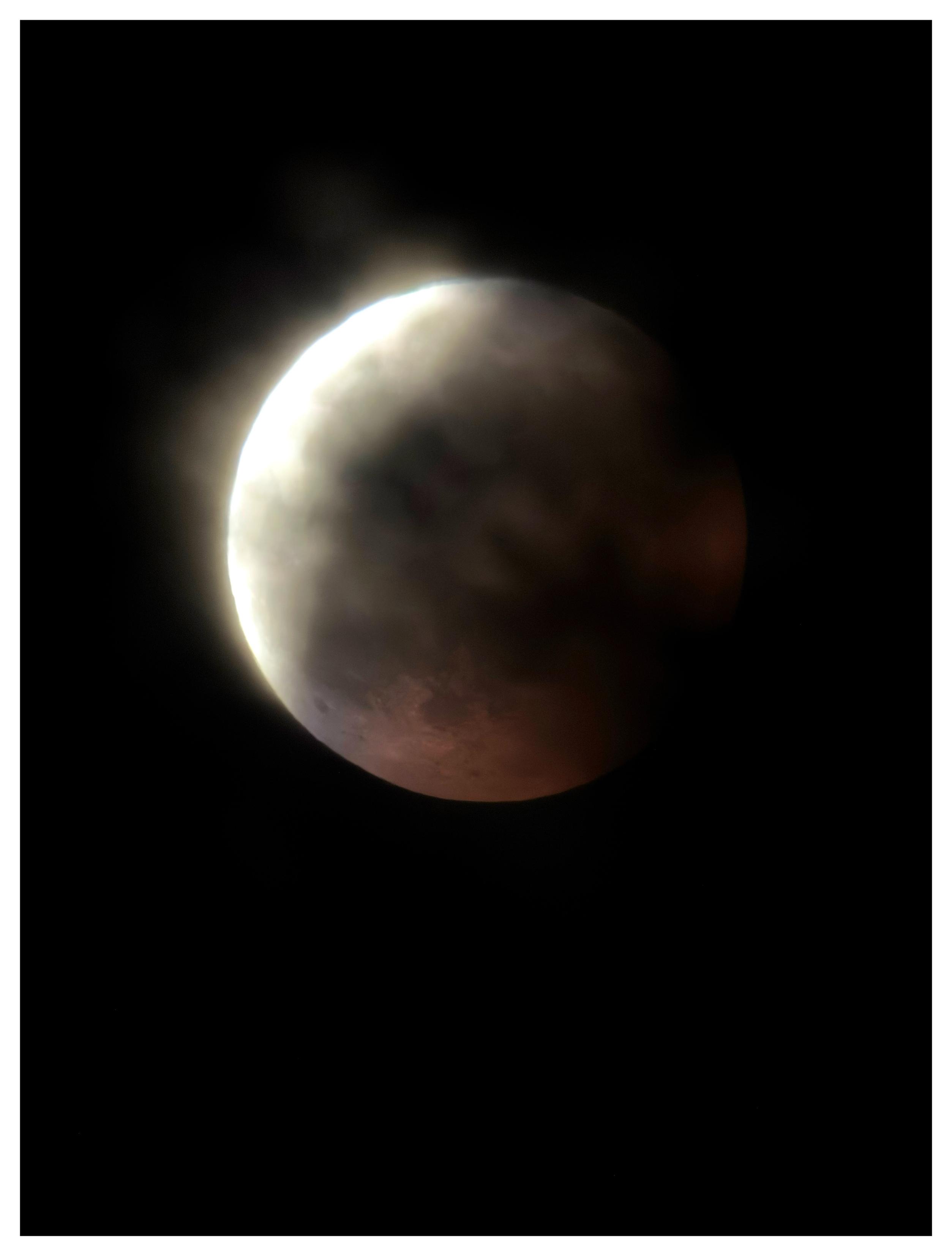 The same lunar eclipse as in the previous photo, but with the moon more in focus and the pine branches blurred into vague dark shadows. The white crescent is overexposed, with a smudged curved edge, while the red surface in Earth’s umbra shows a few crater details.
