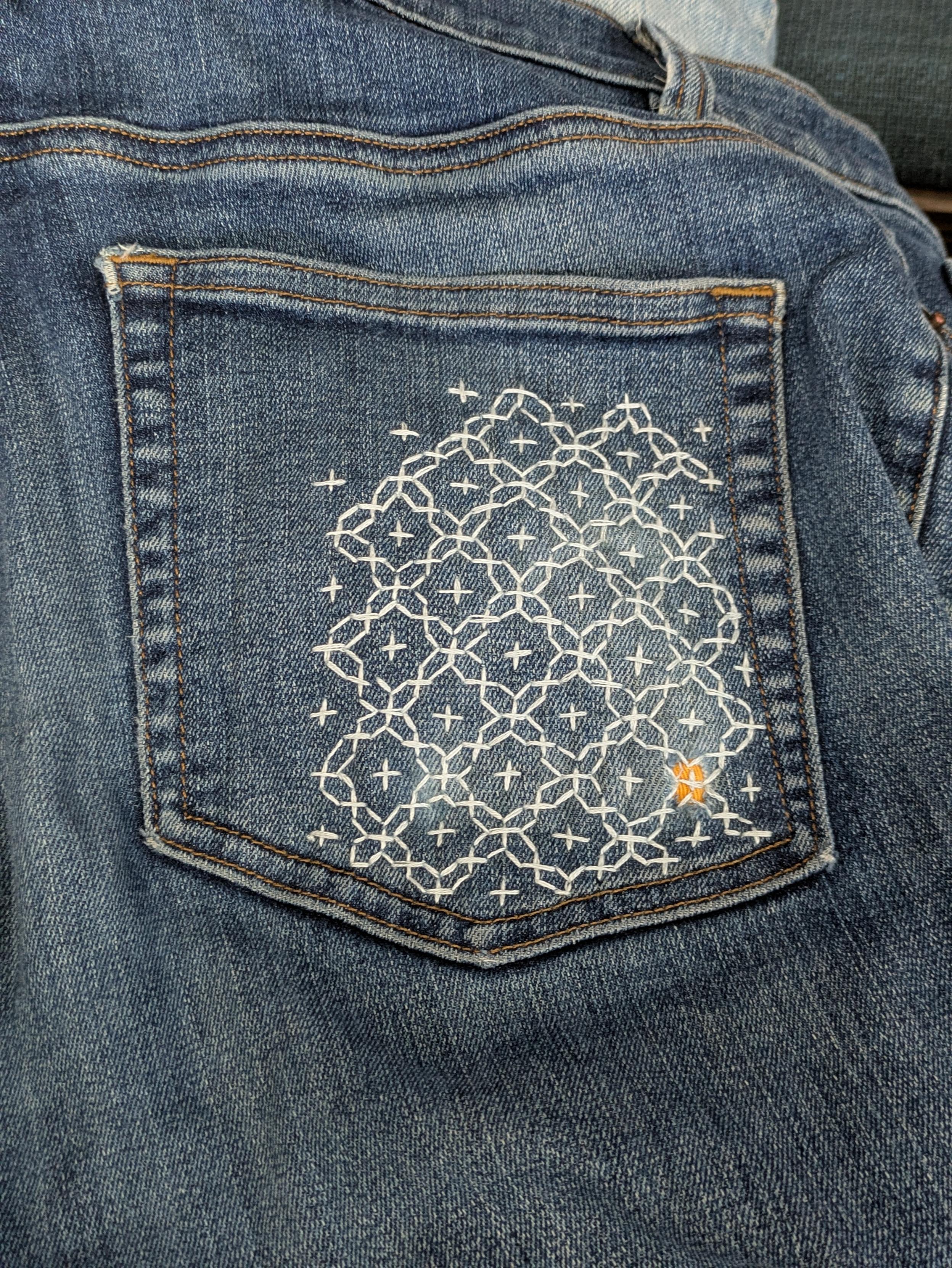 Jeans pocket with hole in lower right corner that is patched from behind with yellow fabric and embroidered down with light blue thread using a sashiko pattern that looks a little like a net and stars