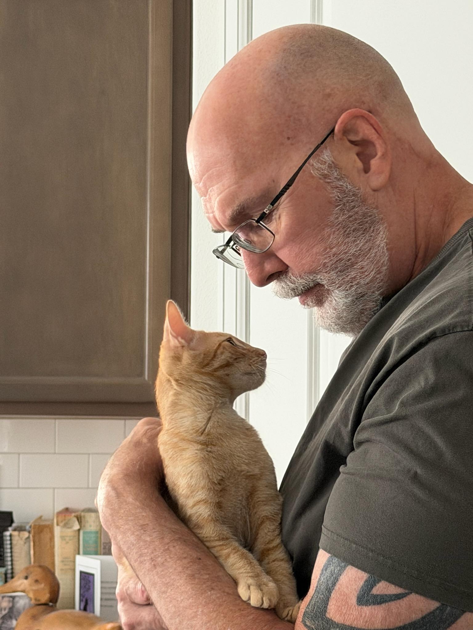 In a Craig, a large man with buzzed head, arm tattoo, beard, and glasses, holds 2-month-old orange kitten Bob close to his chest. Bob has one paw on Craig's arm and is looking trustingly up at his human, who is looking down at him adoringly.
