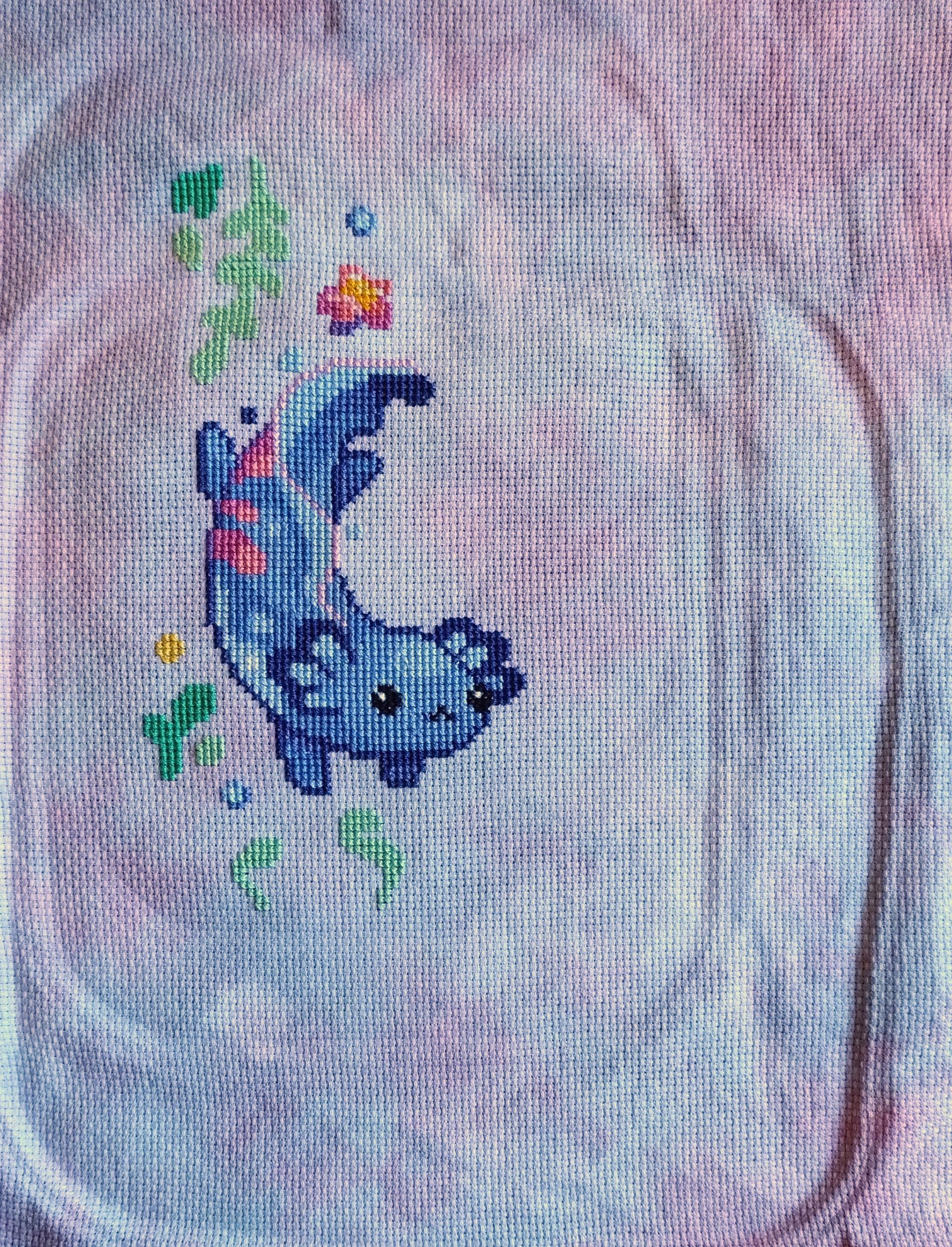 Cross stitch project of a blue and pink axolotl. It is stitched on a grey and pink mottled aida.