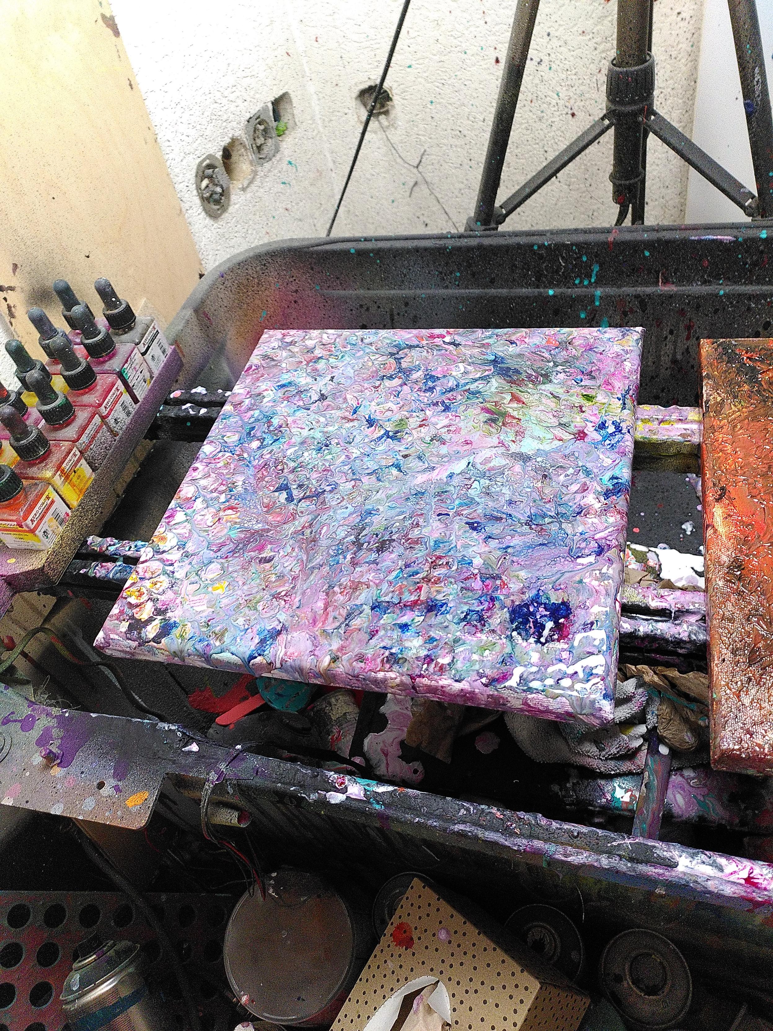 a canvas with wet paint with abstract art in blue, red, yellows in a lot of small cells. Those cells are seemingly random but somewhat form an even pattern. Towards the edges spots of white appears as it was eroding slowly from the edges