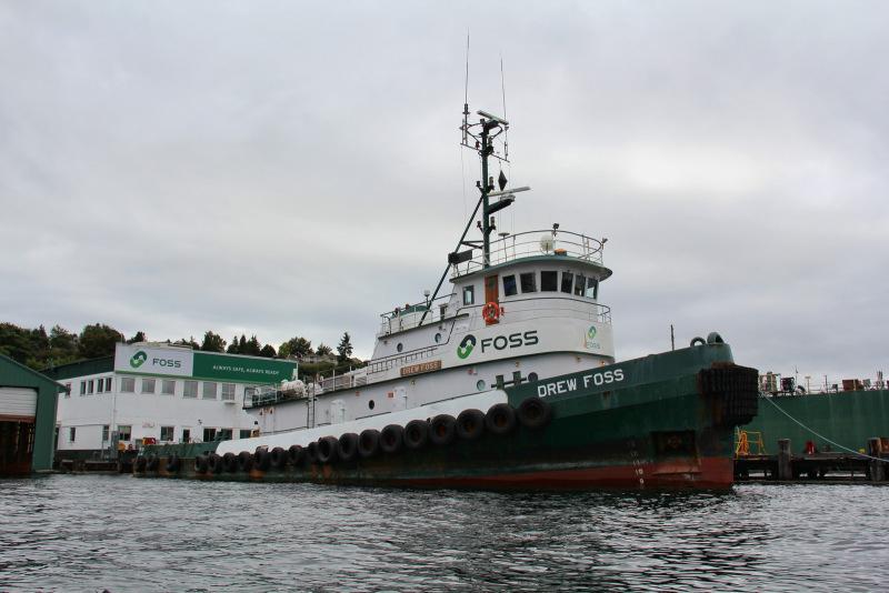 A large tugboat tied to a pier. The hull is green and the structure is white with the word FOSS in large green letters. There is a white building in the background with FOSS on it too.