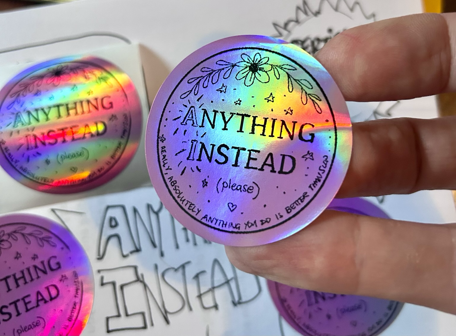 A pink holographic circular label that says “Anything Instead (please), really, absolutely anything you do is better than this slop”