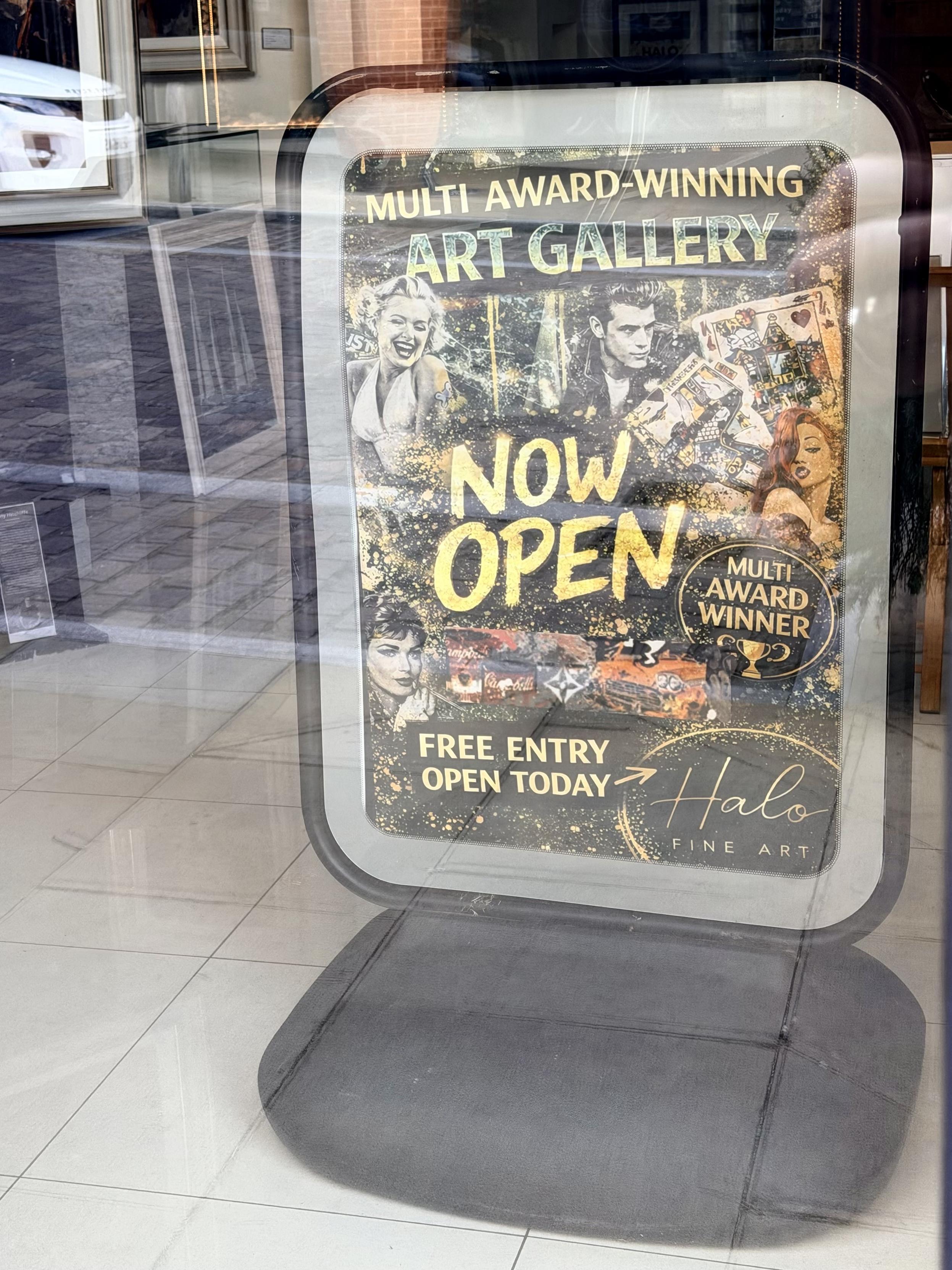 This image features a promotional sign for Halo Fine Art displayed inside a glass window. The sign reads "Multi Award-Winning Art Gallery, Now Open" in large yellow lettering, with "Free Entry Open Today" below it. The background of the sign includes various artistic portraits and a classic orange sports car.