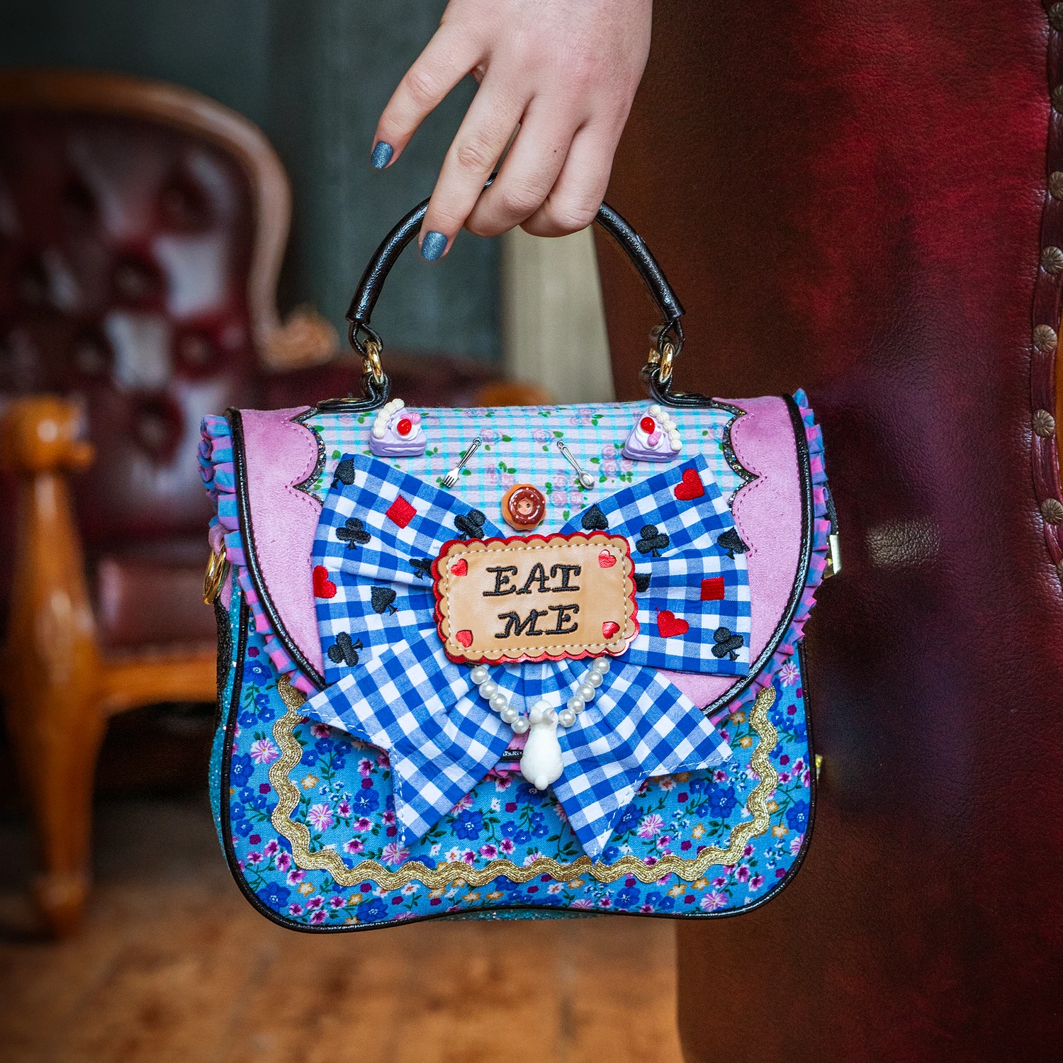 A blue handbag with a patterned floral backsplash, and the cover-flap has a large blue and white chequered bow that says "EAT ME" in the middle, surrounded by hearts.
