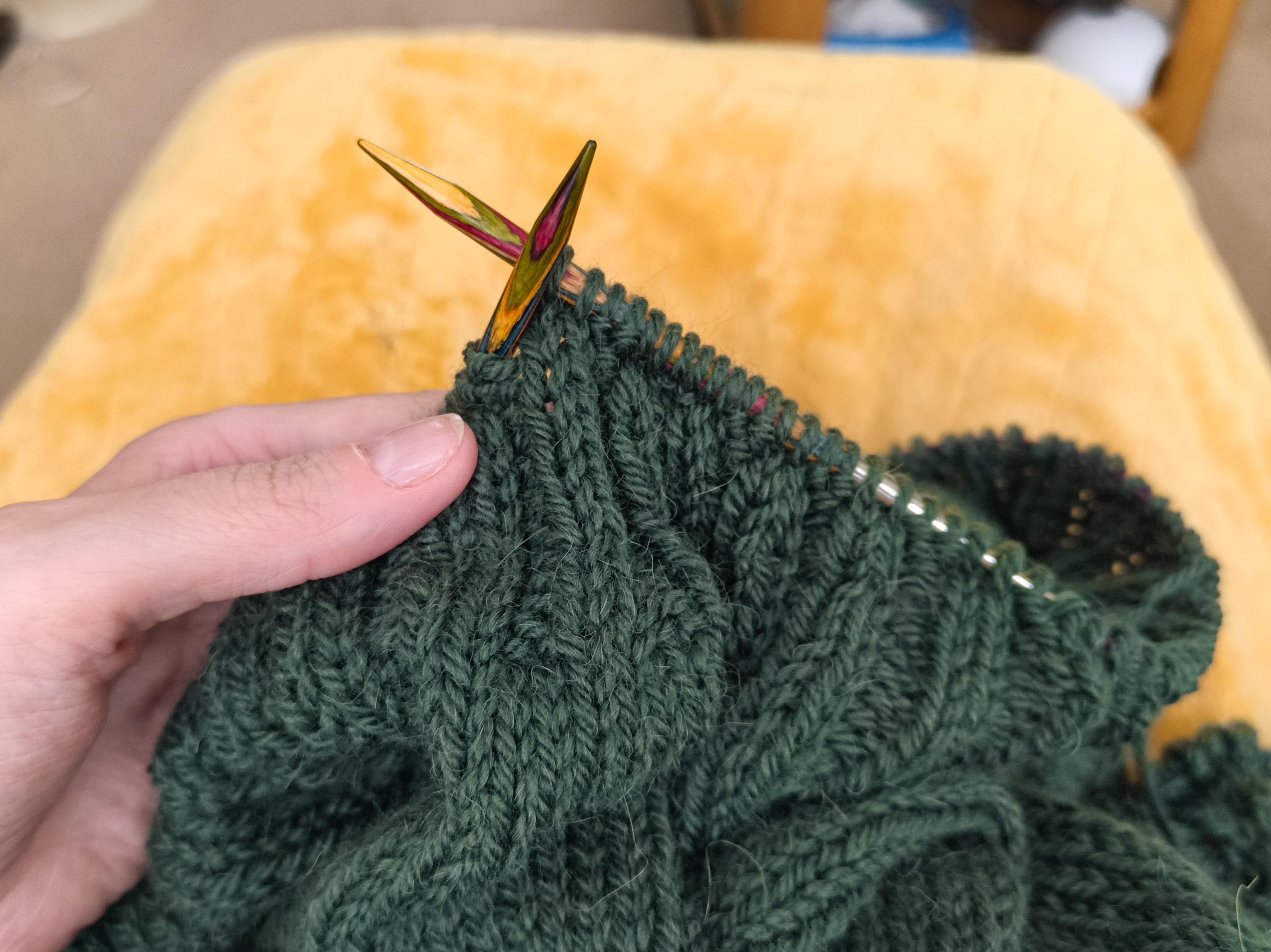 A dark green knitted jumper project on wooden needles, held up in front of a yellow blanket in the background. The visible bit of jumper is mostly plain stockinette with a ribbed edging currently being worked.
