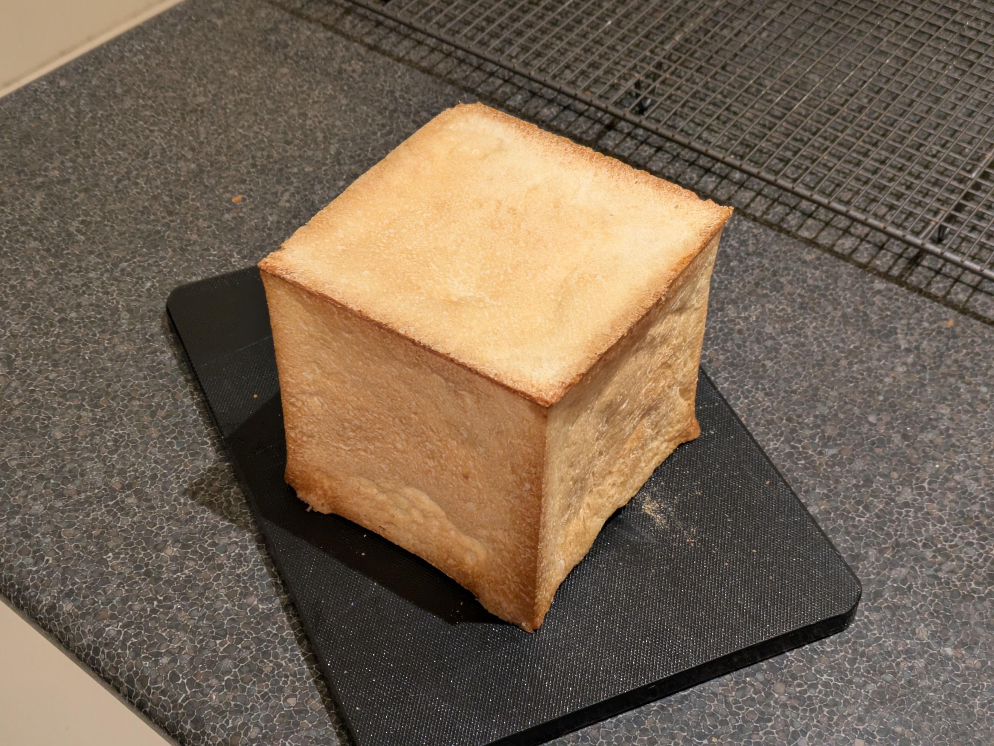 A photo of a cubic loaf of bread sitting on a kitchen cutting board, near a cooling rack. It is an oddly perfect cube with light golden sides and darker brown edges and corners.