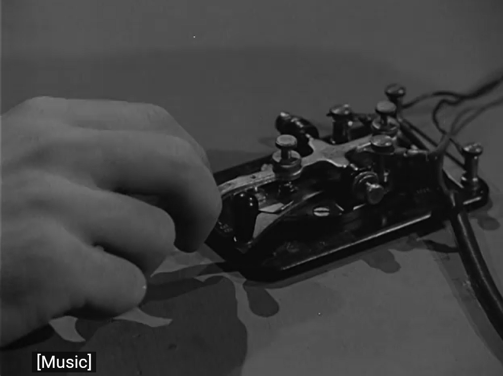 Hand on a strait key tapping out Morse code. The closed captioning reads: [music]