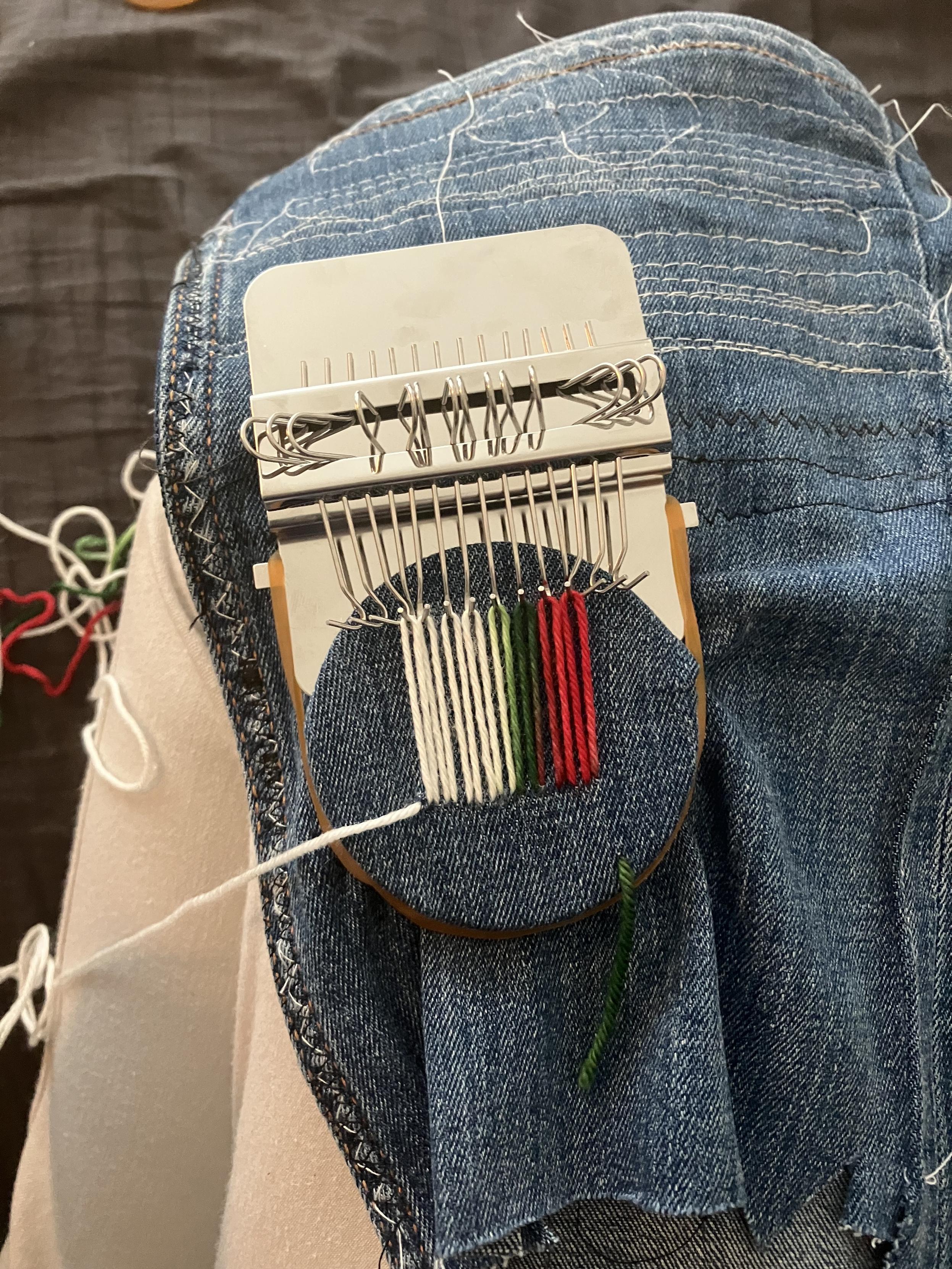 Tiny darning loom on a piece of denim. The fabric has been pulled over a round piece of wood. On top is a metal plate with hooks that can be swiveled 180 degrees. The loom is warped with yarn going through the fabric at the bottom of the patch, up over a hook, back down and through the fabric, etc. It has strands of red, green, and majority white.