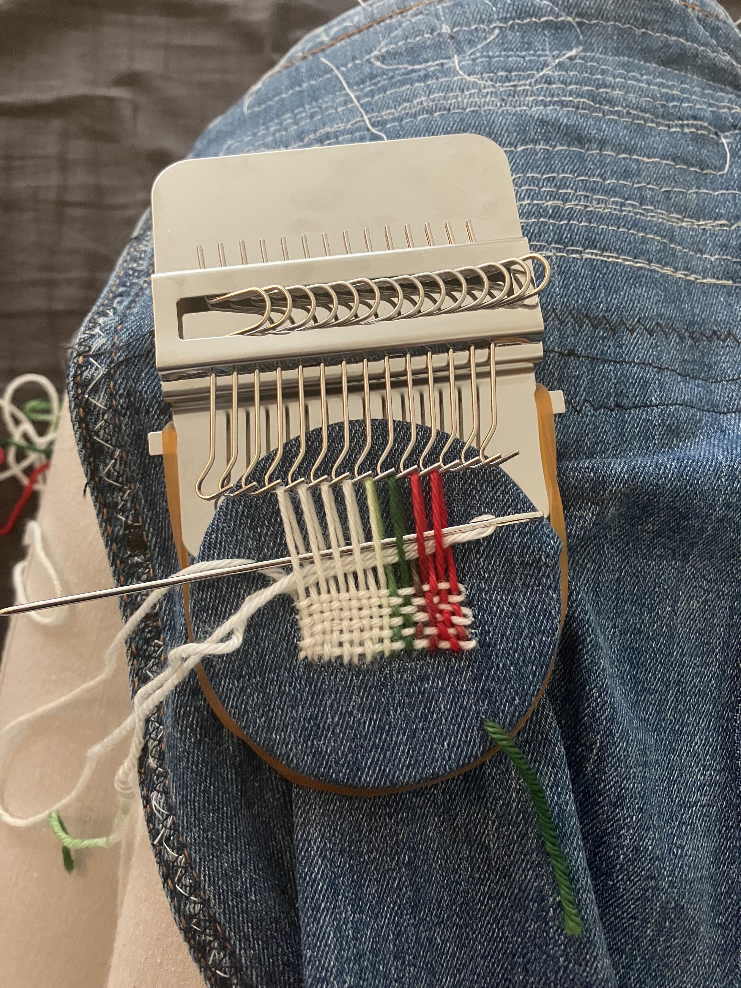 Tiny loom in use. Several white strands have been woven through horizontally and a long needle is just about to add a new one.
