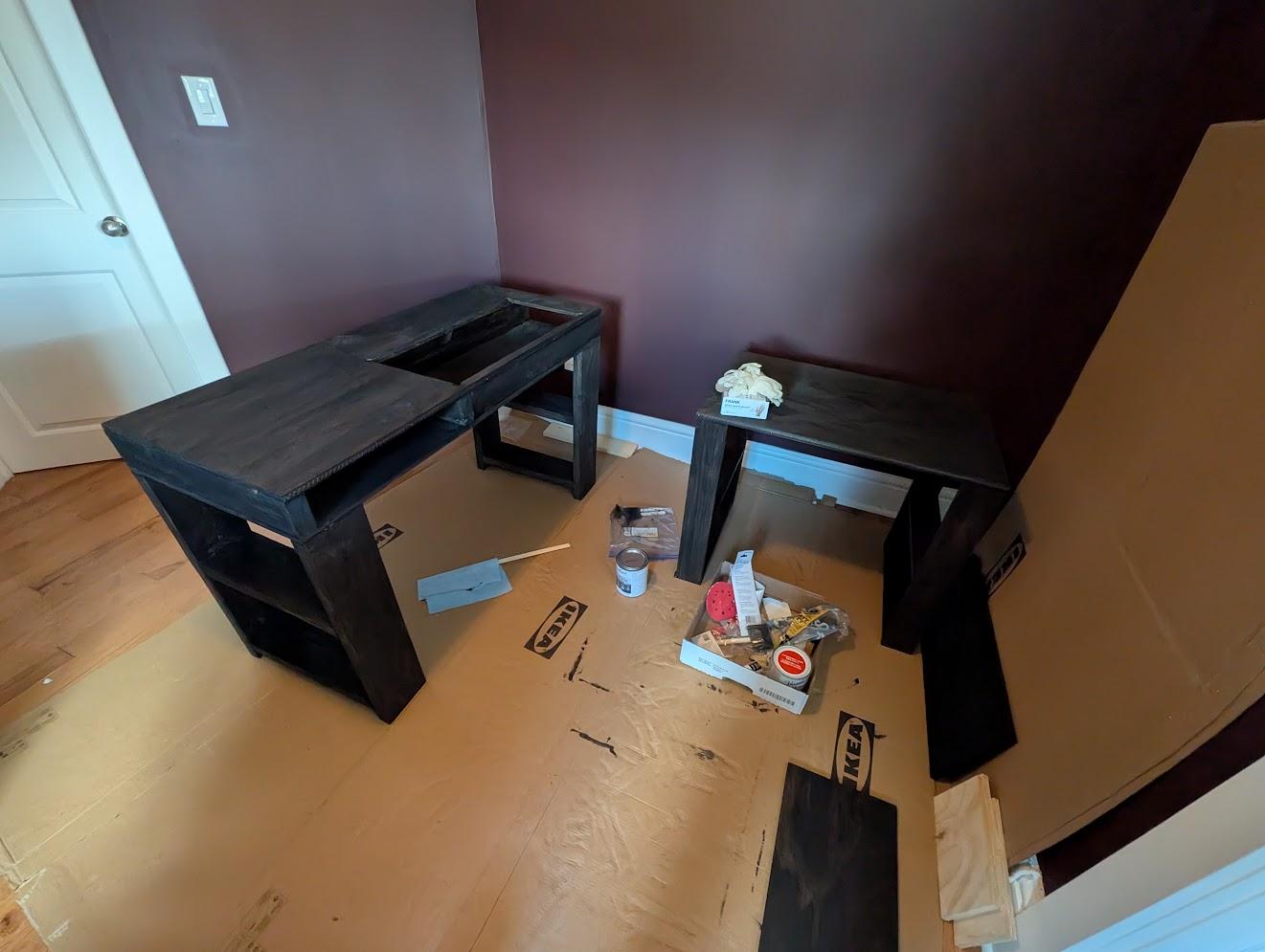 Two sewing tables with dark stain sitting on top of cardboard amidst staining/varnishing supplies