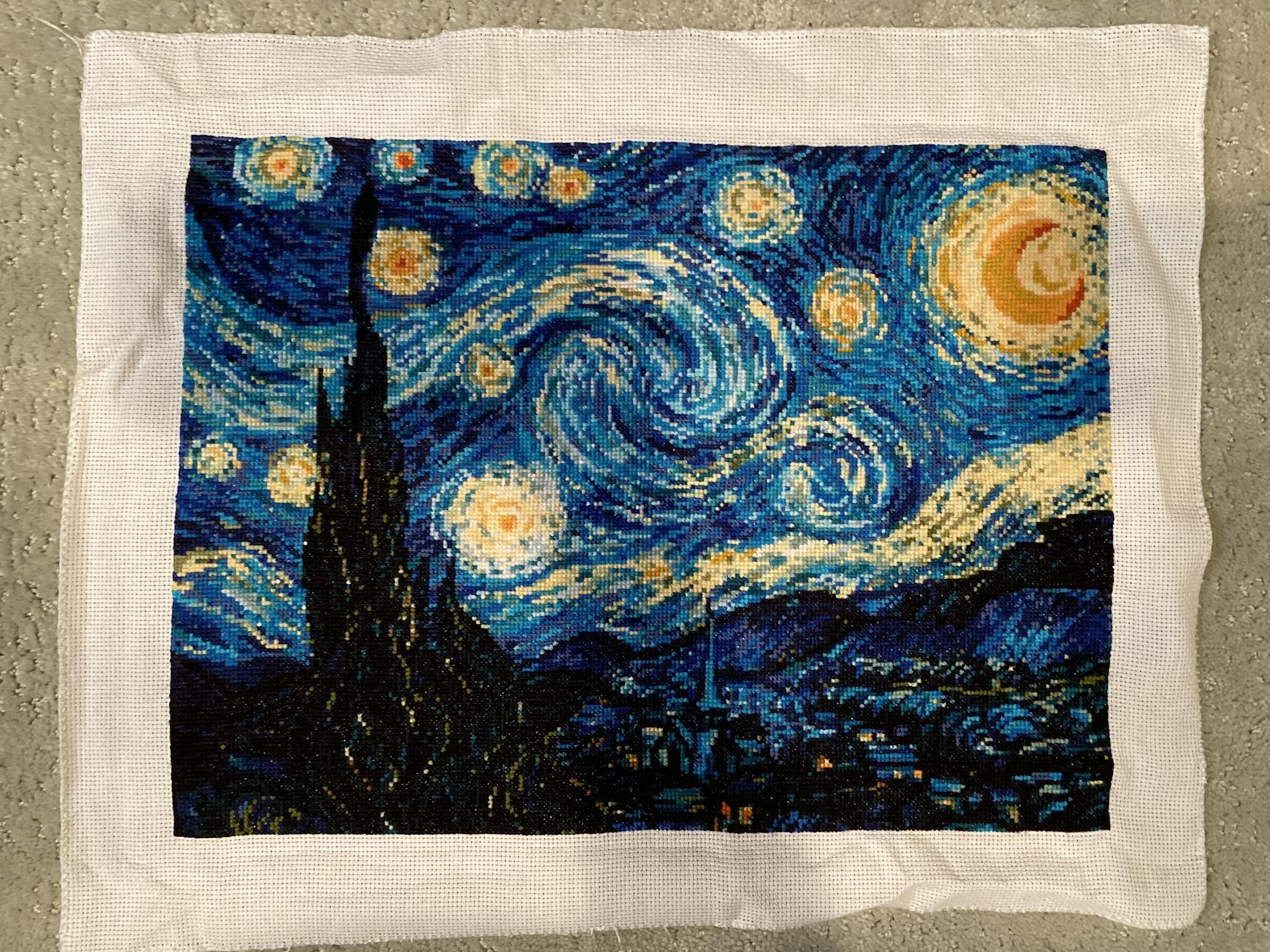 A rectangular piece of blue, yellow, and white cross-stitch embroidery is centered on a larger piece of white canvas. The embroidery features a swirling night sky filled with various-sized bright yellow and white circular shapes. A large, glowing yellow crescent moon is visible in the upper right corner. A tall, dark, jagged silhouette stands on the left side, and a dark, textured landscape with small points of light occupies the bottom of the piece. The entire embroidered work is surrounded by a wide, plain white fabric border.
