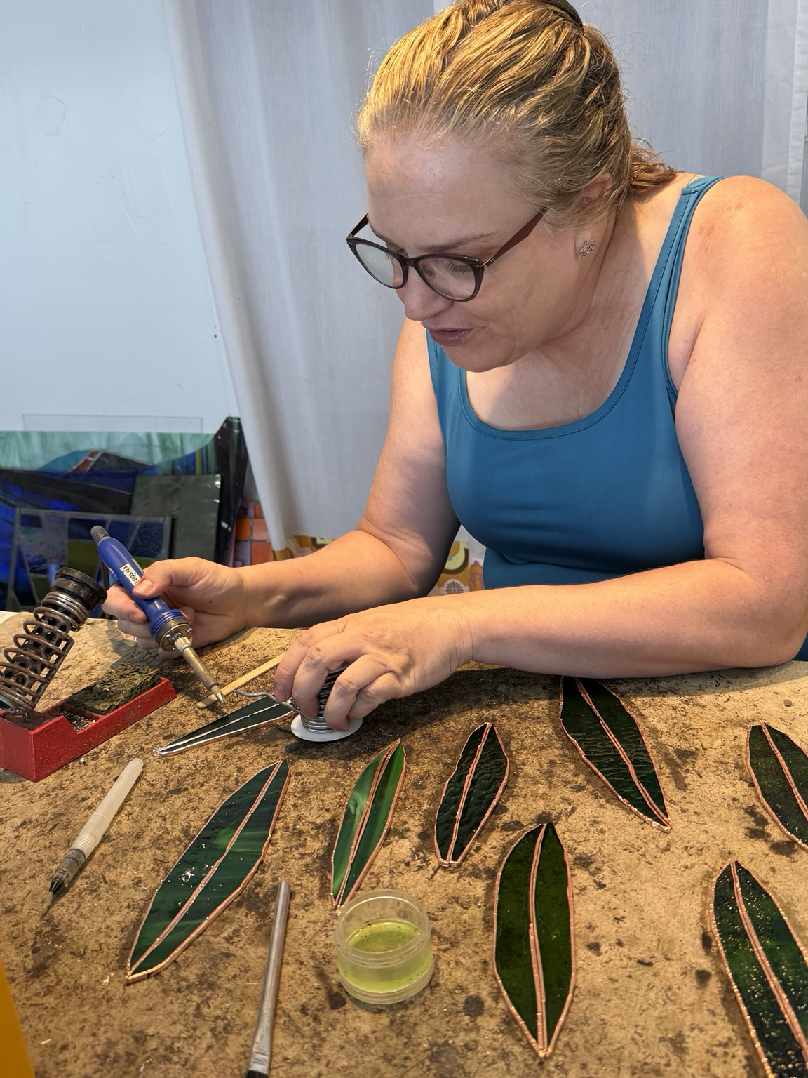 A white woman with glasses and a blue dress is focused on soldering some green glass