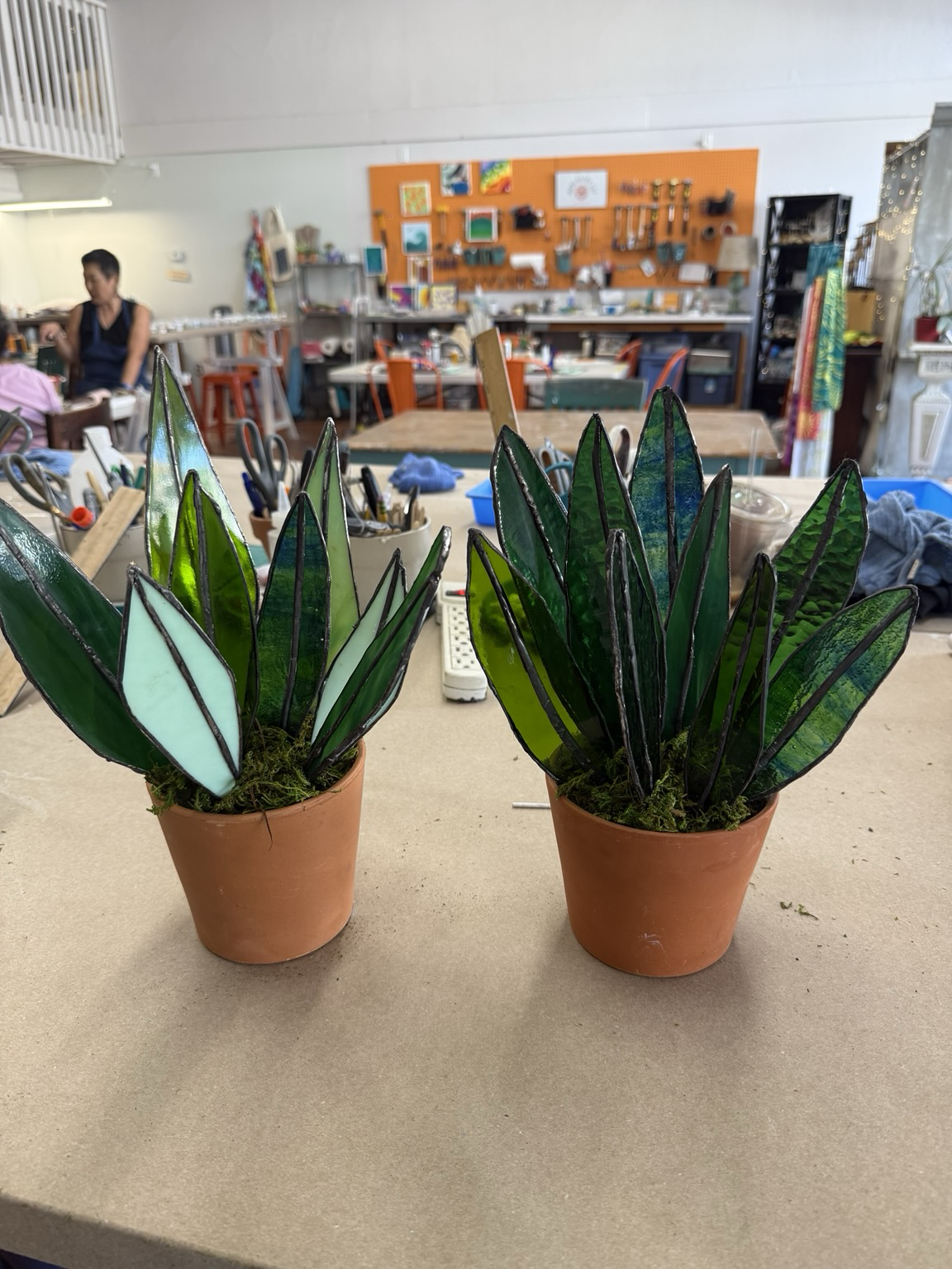 Two potted plants made of stained glass, featuring green and light-colored leaves, are displayed on a workbench. In the background, a crafting workshop is visible, with various tools and artwork.