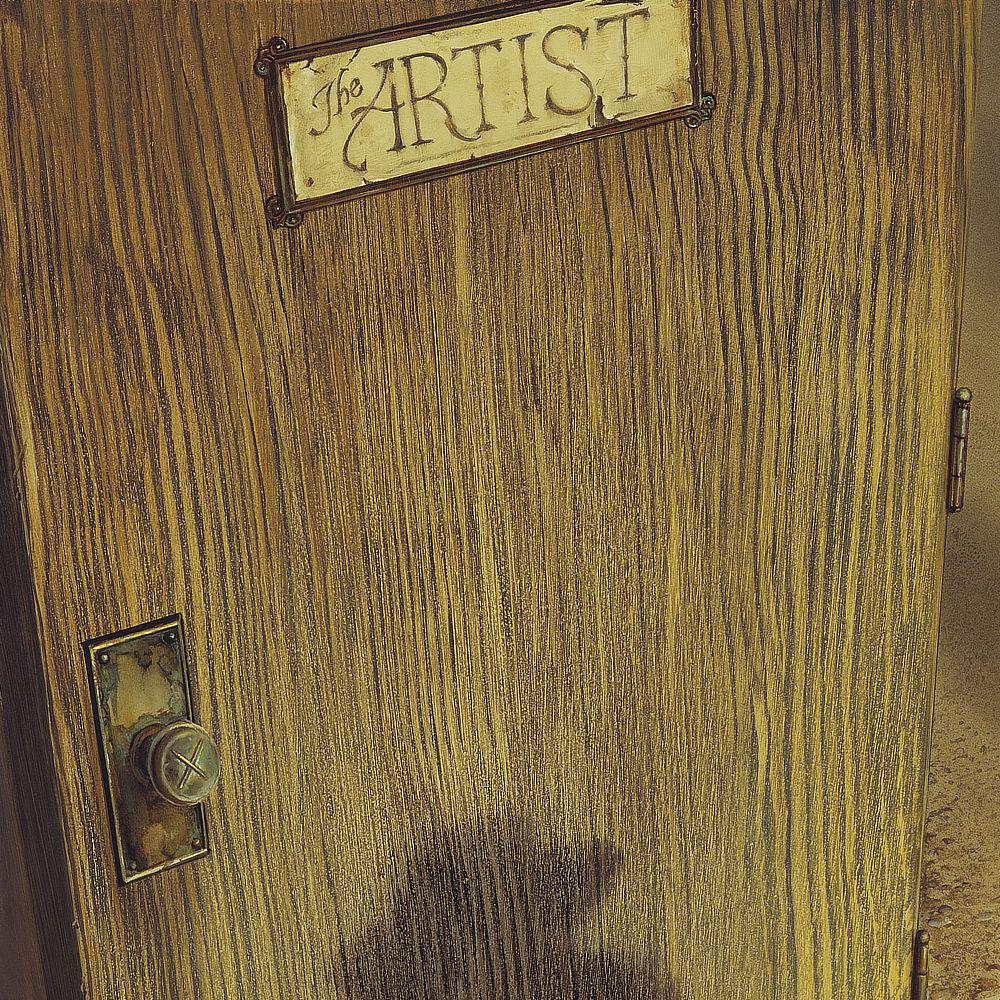 Close detail of the yellow and brown woodgrain of a door. At the top is a sign with cracking paint that reads”The ARTIST.” On the lower left a doorknob marked with the X of two crossing paint brushes sits on a metal plate rusted and peeling.