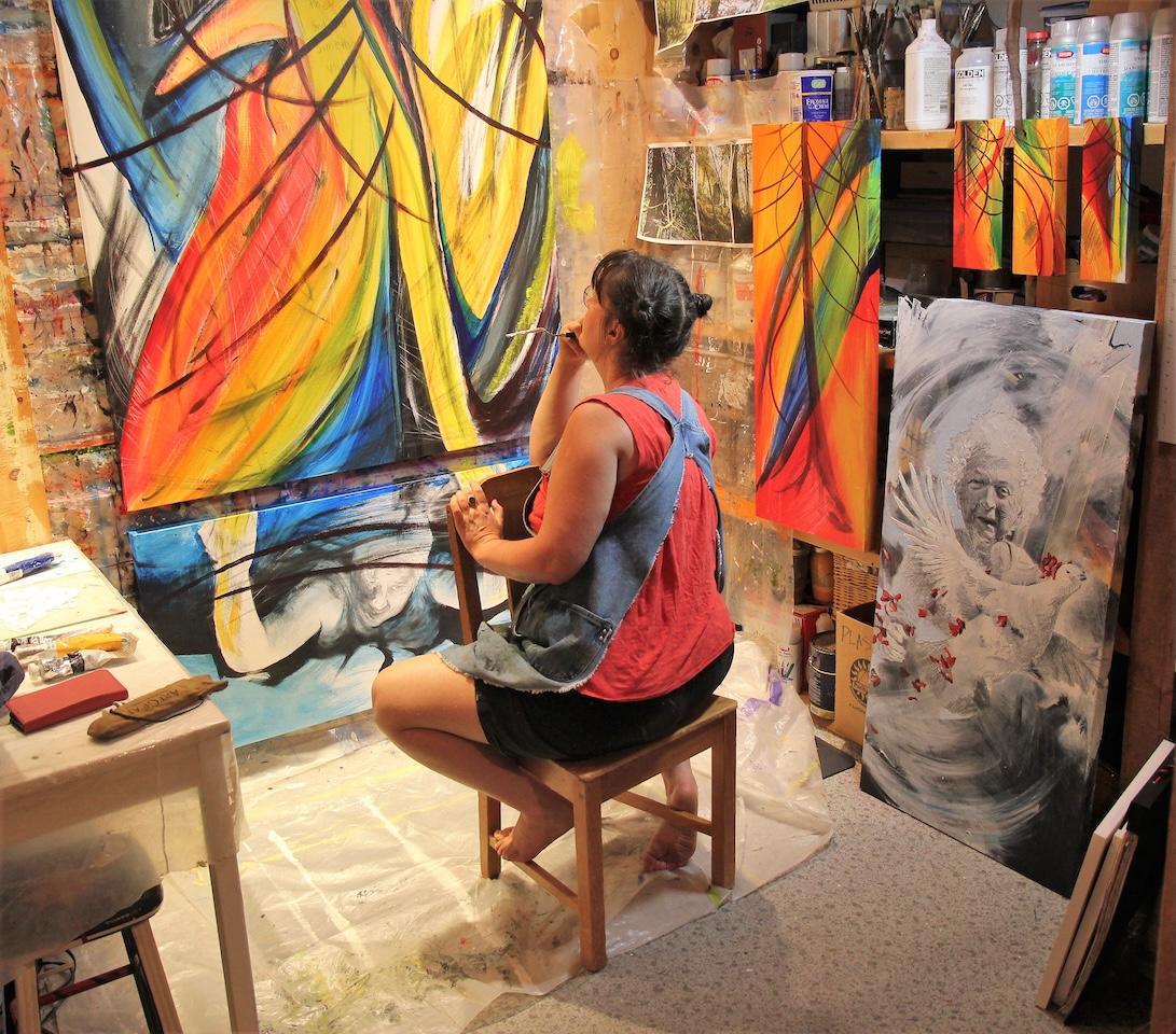 Canadian eco-social practice artist Emily van Lidth de Jeude in her studio, 2023, working on a series about wildfires. Beside her on the floor stands a portrait of her Ukrainian grandmother.