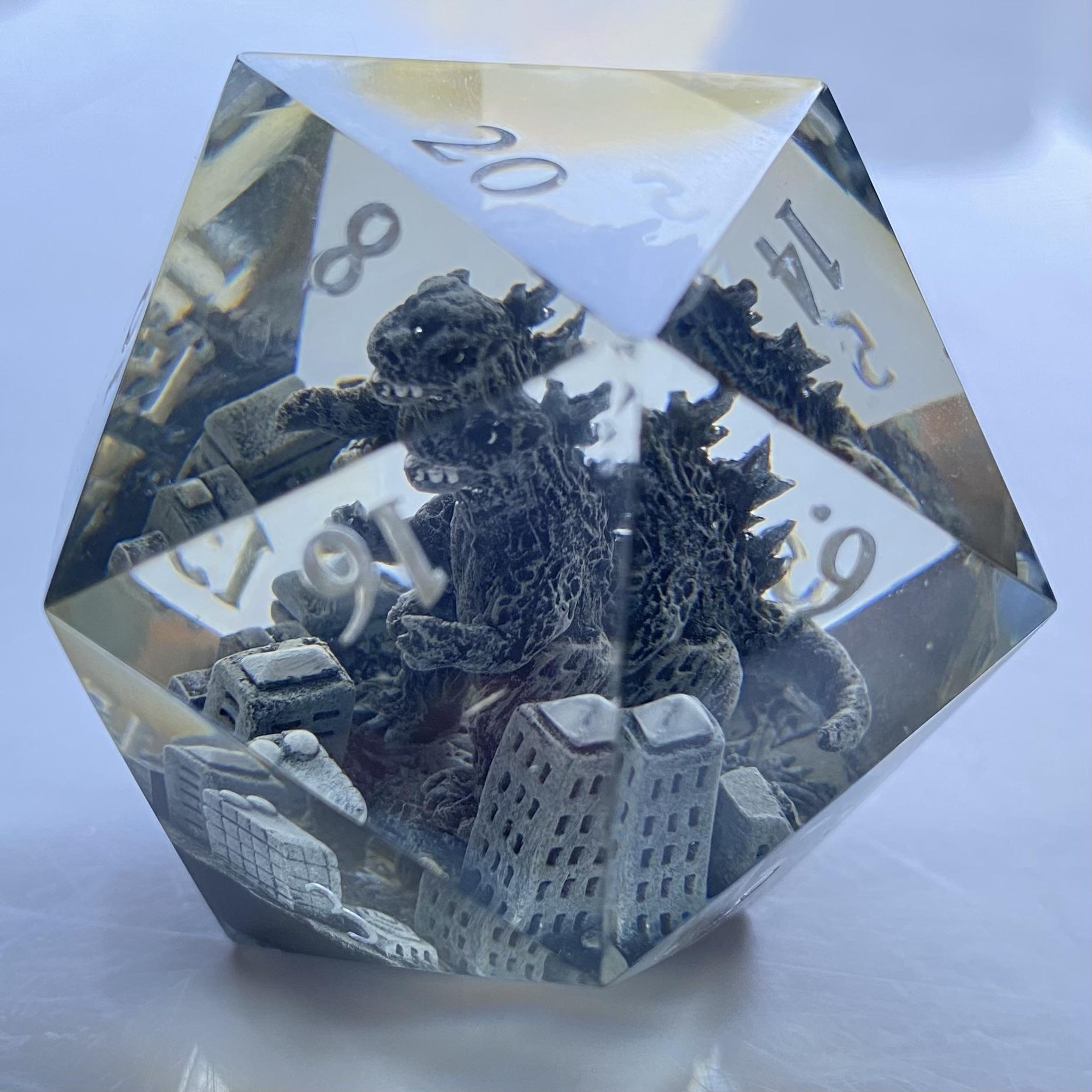 A monotone Godzilla destroys a monotone city, inside a clear resin d20. The numbers are painted in pearly white