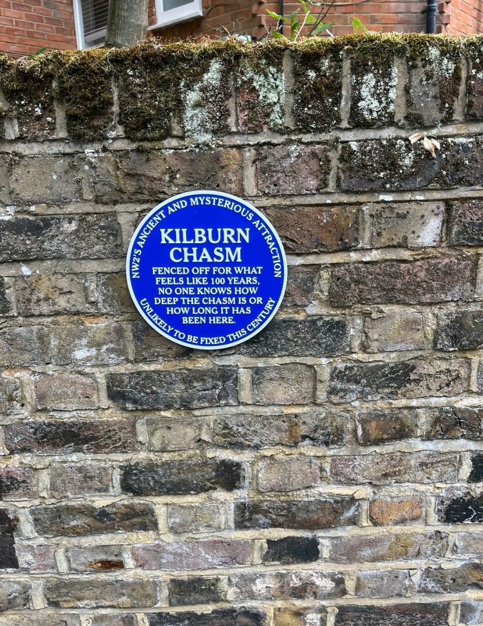 A convincing looking but fake blue plaque on a wall. Text reads: "Kilburn Chasm. NW2's ancient and mysterious attraction. Fenced off for what feels like 100 years. No one knows how deep the chasm is or how long it has been here. Unlikely to be fixed this century"