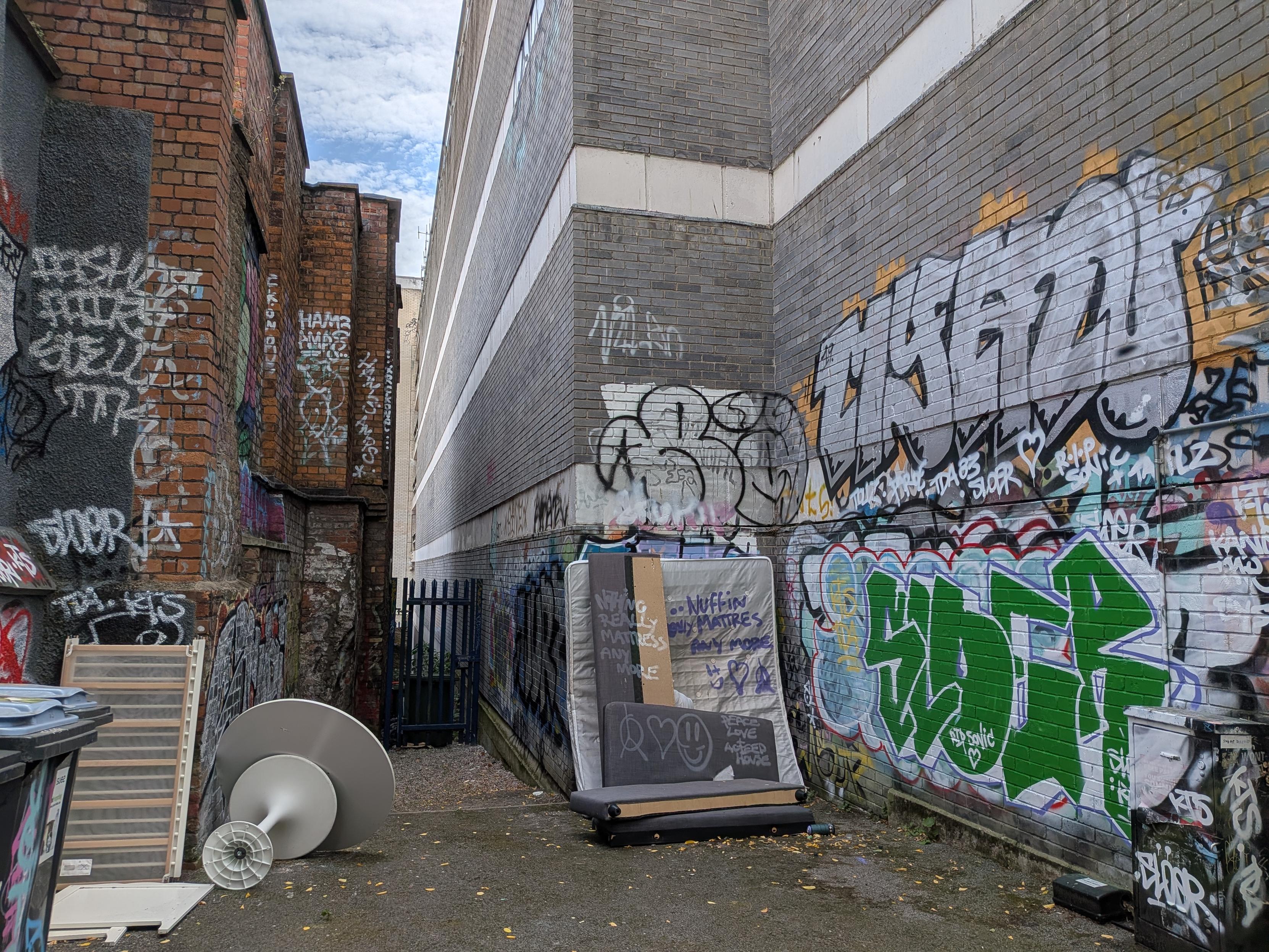 A dank alleyway between two buildings where some broken furniture has been left. The walls are covered in huge graffiti tags. A mattress and some broken up sofa parts lean up against a wall with some text sprayed onto them.