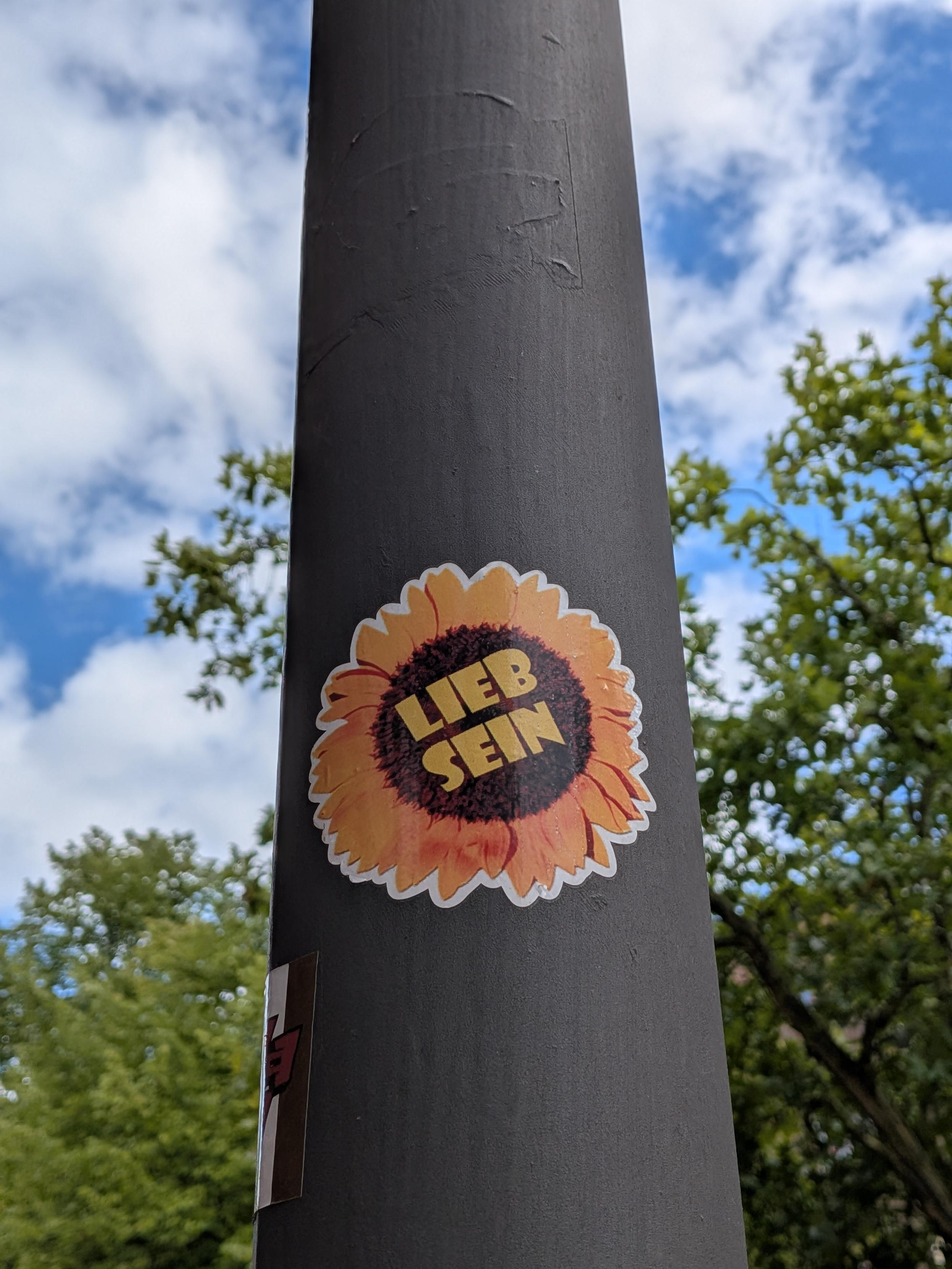 Sticker on a lamppost, a sunflower with "Lieb sein" (be nice, or lovely) in the middle.