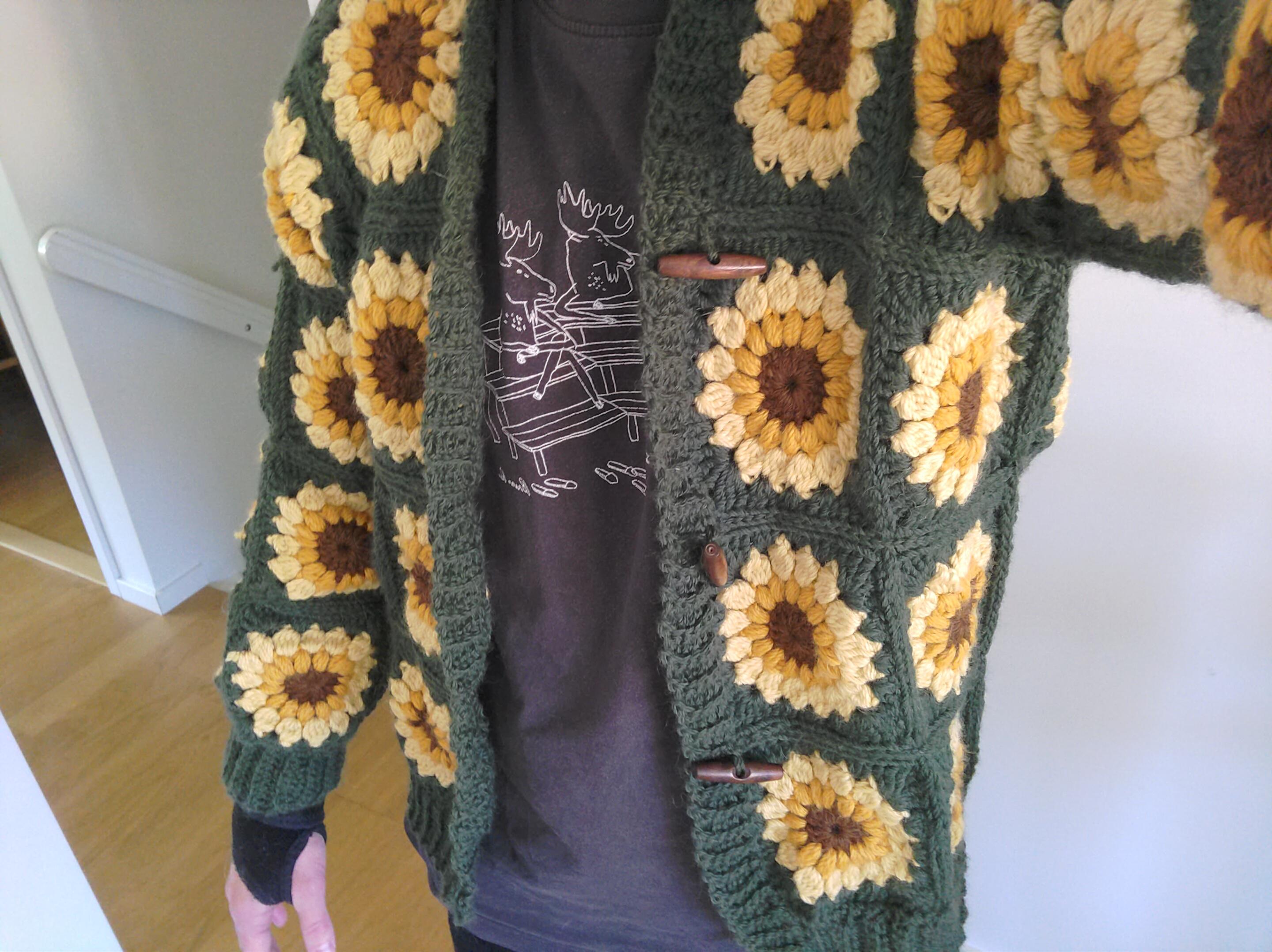 My torso sporting a crocheted cardigan made of granny squares with a pattern of sunflowers surrounded by ivy green yarn.