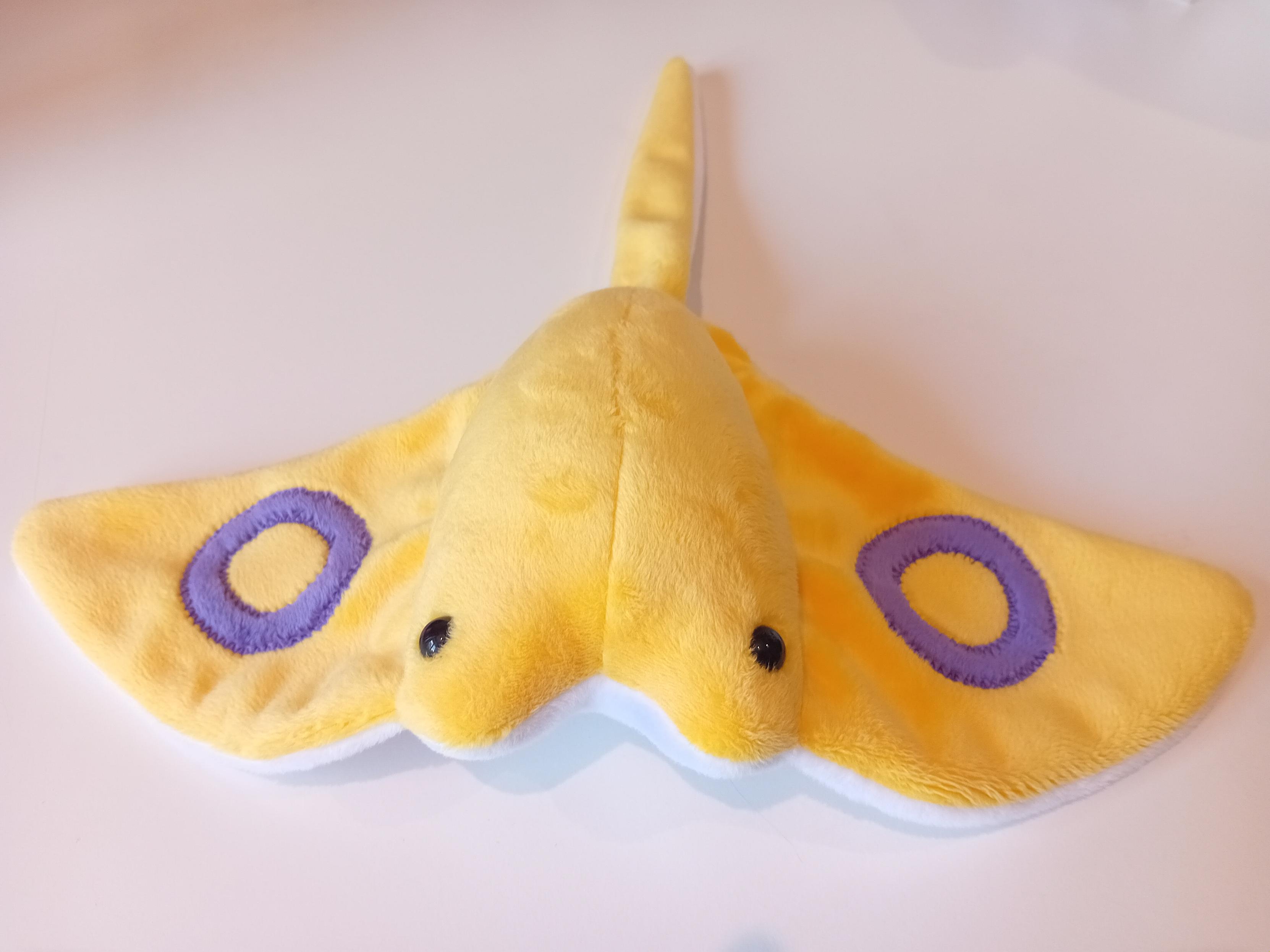 An intersex flag Seaflap (stingray) plushie. They're sunshine yellow with two purple rings, one on each wing. They have a white belly.