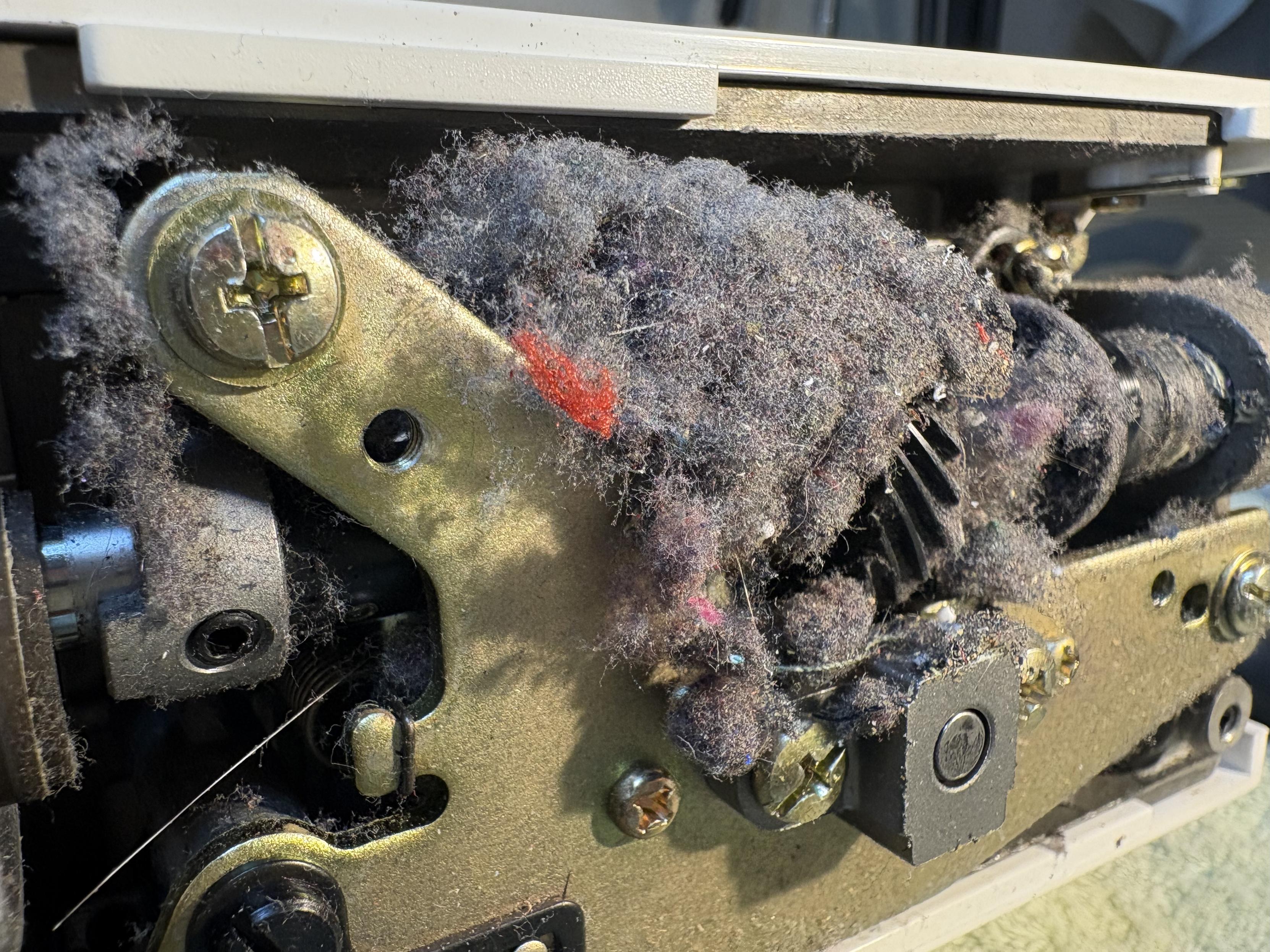 The internals underneath the needle plate & bobbin area of a sewing machine. It's overflowing with lint. The lint is getting caught in the gears.