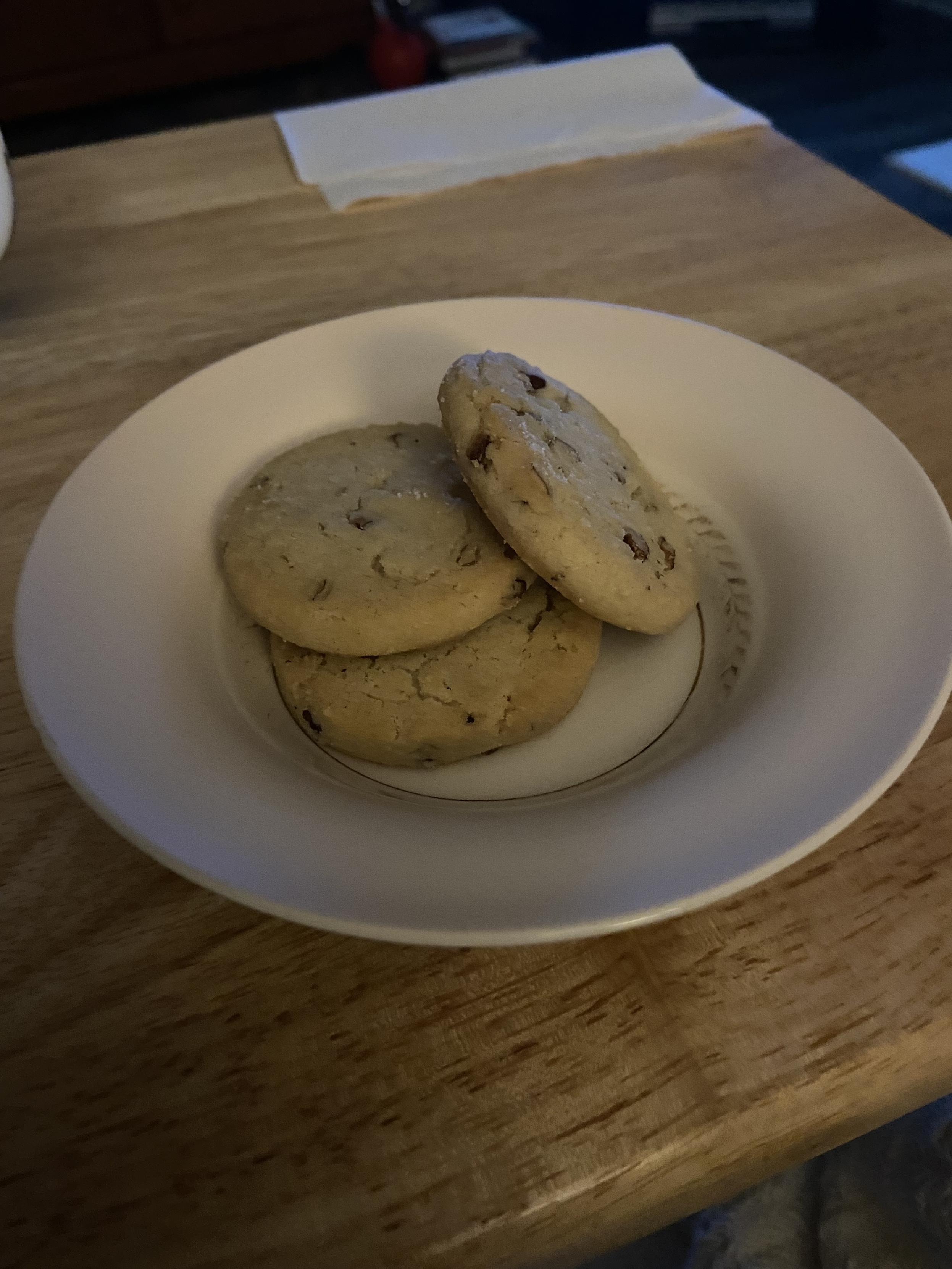 A little pink bowl contains three cookies. The bowl is sitting on a wooden table. A white napkin is in the corner of the table.